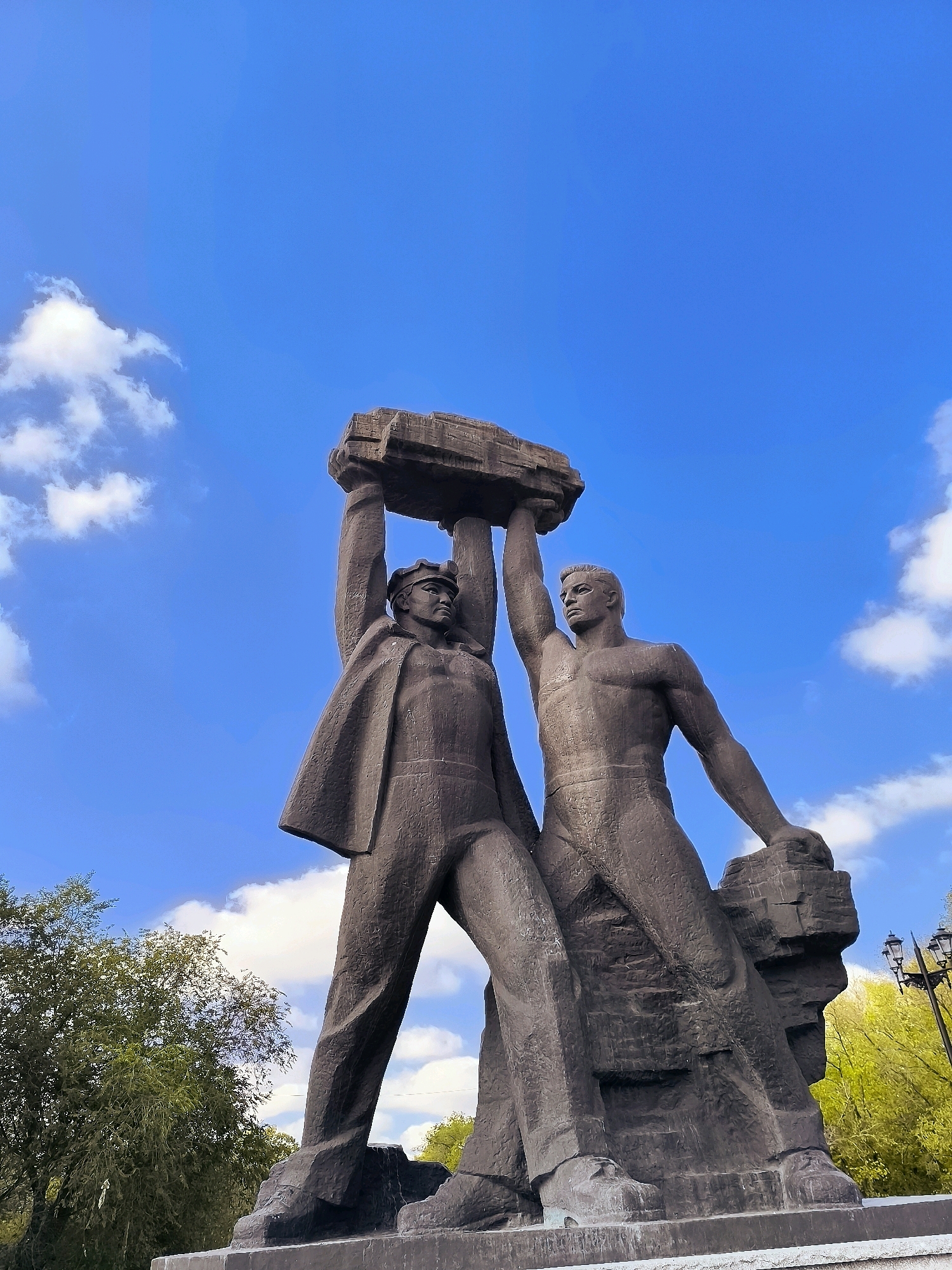 This is a low-angle, full-shot photograph of a large, dark-colored stone monument depicting two muscular men raising a block of stone above their heads. The monument is set against a bright blue sky with scattered white clouds. The man on the left is wearing a cap and a coat draped over his shoulders, while the man on the right is bare-chested and wearing pants. Both men appear to be straining to lift the heavy stone block. In the background, trees with green and yellow foliage are visible, along with a lamppost. The monument is situated outdoors, likely in a park or public square.