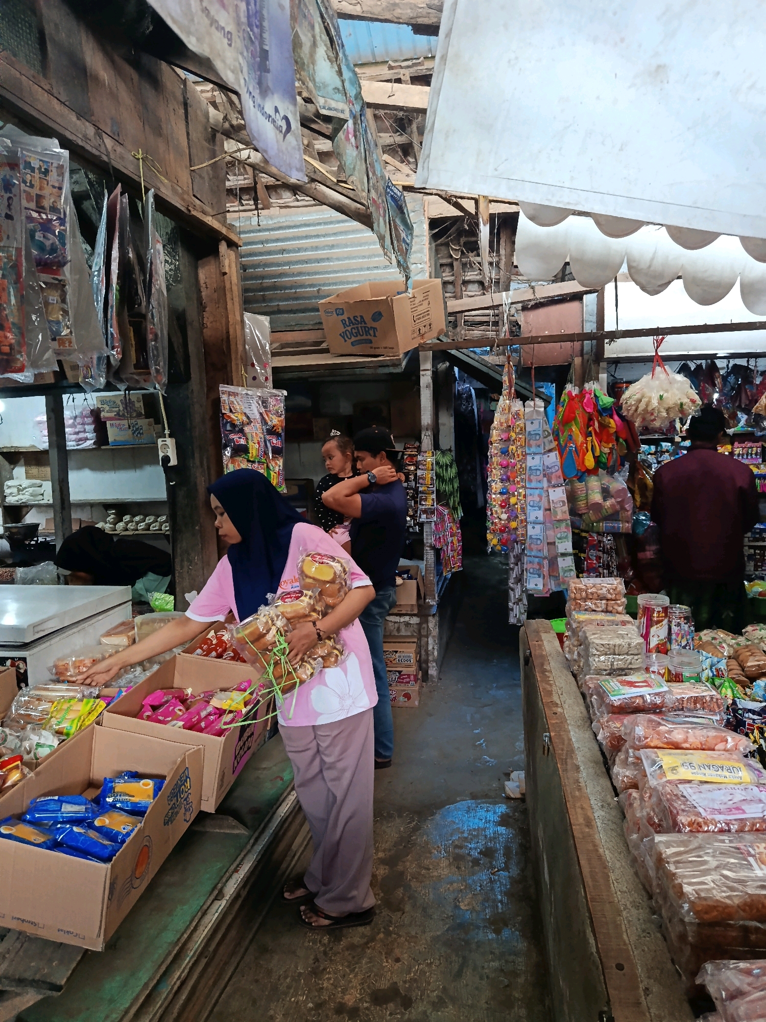 The image shows a bustling market scene in Mlawang, Indonesia. In the foreground, a woman wearing a blue hijab and a pink shirt is reaching into a cardboard box filled with blue and yellow packaged snacks. She is holding a bunch of packaged baked goods tied with green string in her right arm. To her left, another cardboard box contains similar packaged snacks. Behind her, a man is holding a young child. The market stalls are made of wood and metal, with various goods displayed, including packaged snacks, dried goods, and colorful balloons. The floor appears to be concrete, with some puddles of water. The overall impression is of a vibrant and active marketplace.