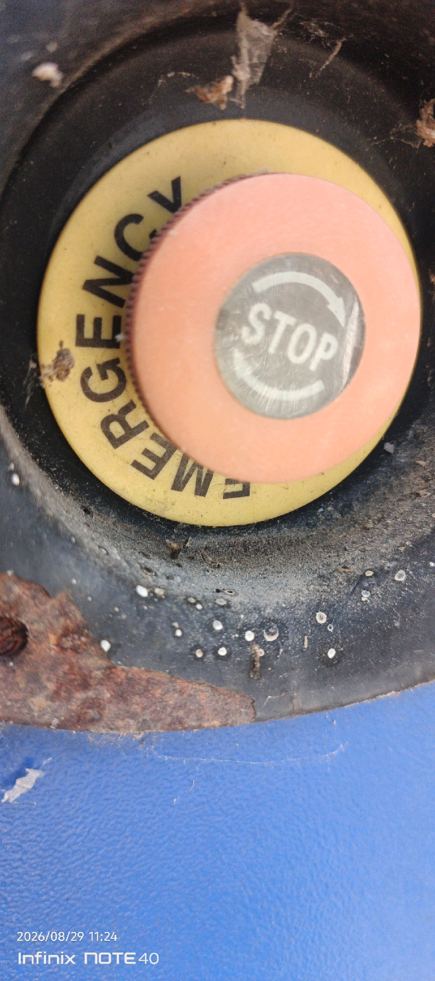 The image shows a close-up of an emergency stop button on what appears to be industrial equipment. The button itself is round and orange, with a silver circular sticker in the center displaying the word "STOP" in white capital letters, enclosed by two curved arrows. Below the orange button, a yellow circular label is visible, with the word "EMERGENCY" printed in black capital letters. The yellow label is partially obscured by the orange button. The surrounding area of the equipment is dark and appears to be made of metal, with some signs of rust and dirt. The bottom portion of the image shows a textured blue surface, likely part of the machinery. In the bottom left corner, text indicates a date and time "2026/08/29 11:24" and a device name "Infinix NOTE 40". The image does not depict any people, actions, or interactions, nor does it provide any visual information that would allow for the identification of a specific location in the city of Patampanua, Indonesia.