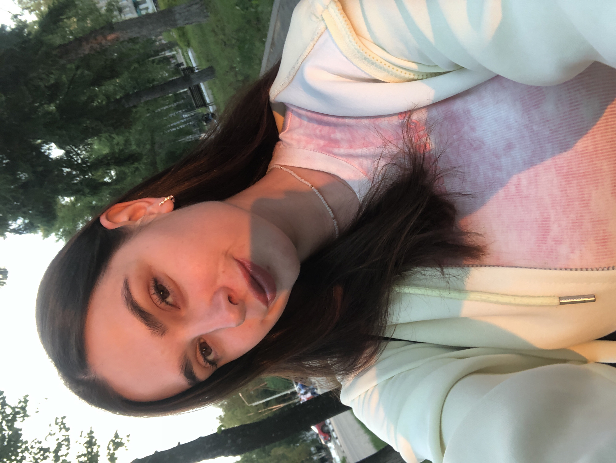 A young woman with long, dark brown hair, wearing a white and pale yellow zippered hoodie over a pink tie-dye t-shirt and a beaded necklace, is pictured in an outdoor setting. She is looking towards the camera with a slight smile. The background features trees, grass, a sidewalk or road, and a red car in the distance, suggesting an urban park or street in the city of Chaikovsky, Russia. The lighting indicates it is either sunrise or sunset, casting a warm glow on the scene.