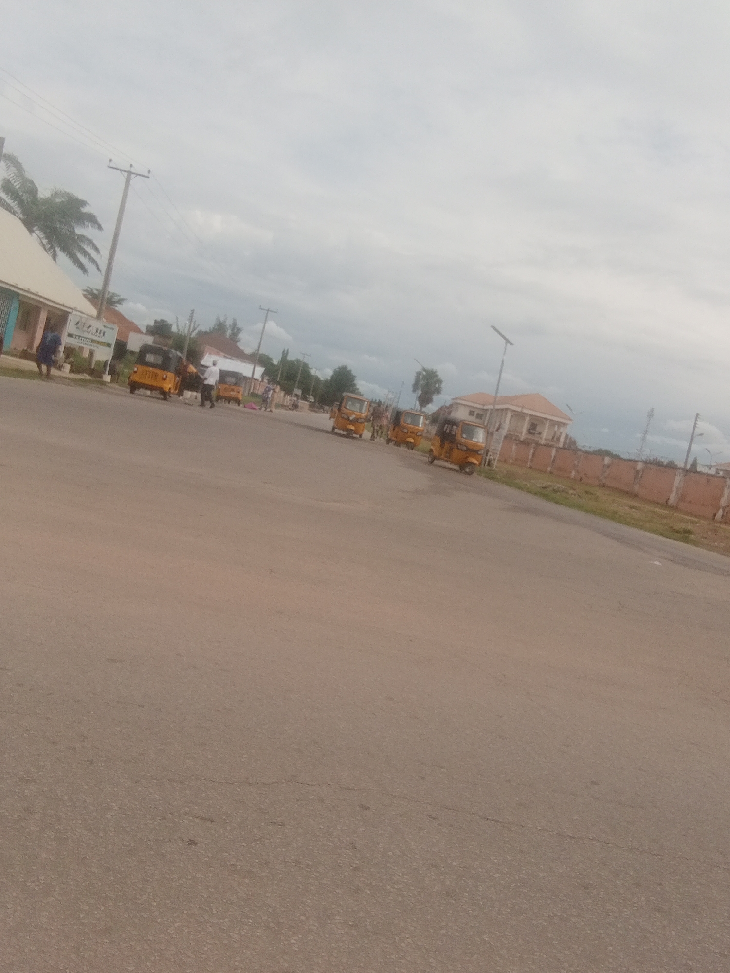 The media file shows a street scene in Jalingo, Nigeria, under an overcast sky. Several yellow auto-rickshaws, commonly known as "Keke Napep," are visible on the road. Some are parked, while others appear to be in motion. A few people are present, some standing near the auto-rickshaws and others walking.
On the left side of the image, there is a building with a light-colored facade and a smaller blue structure attached. A sign is visible in front of the building, but the text is illegible. Palm trees are also present, suggesting a tropical or subtropical climate. In the background, there are more buildings, a fence, and utility poles with overhead wires, indicating a developed area. The road itself is paved and appears to be a main thoroughfare. The overall mood is calm and ordinary, depicting a typical day in a Nigerian town. The time of day seems to be daytime, but the cloudy weather obscures direct sunlight. There are no overt signs of any specific event or heightened activity.