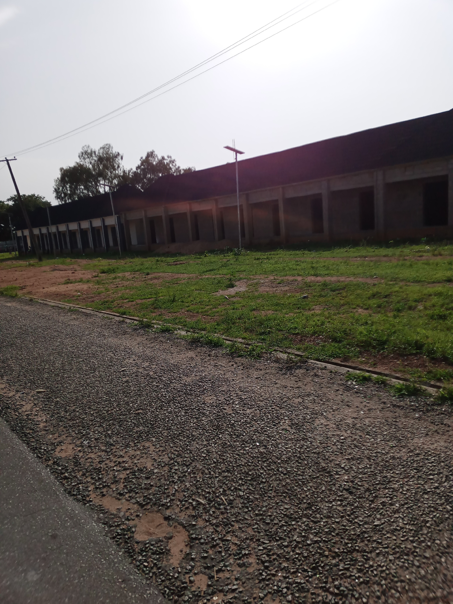 The image depicts an outdoor scene in Jalingo, Nigeria, with a long, low-rise building under construction or renovation. The building has a dark, sloped roof and a series of open, rectangular doorways or window frames along its facade. The exterior walls appear to be made of concrete or brick. In front of the building, there is a strip of uneven ground with patches of dry earth and sparse green grass. A gravel path runs diagonally across the foreground of the image, leading the viewer's eye towards the building. A utility pole with power lines is visible to the left of the building, and a solar-powered street light stands in front of it. The sky is bright and overcast, suggesting a daytime setting. The overall impression is one of an unfinished or abandoned project in a semi-urban or rural environment.