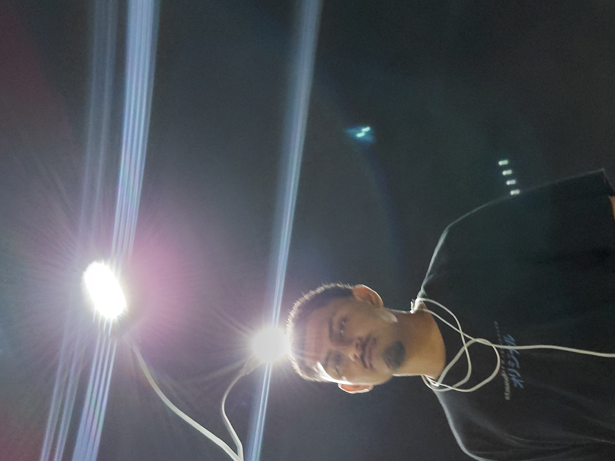 A nighttime, low-angle shot in Santa Ana, Venezuela, captures a male individual positioned against a dark background with significant artificial backlighting. The subject, an adult male with medium skin tone, short dark hair, a goatee, and a thin mustache, looks slightly upwards and to the viewer's left. He wears a dark-colored short-sleeved top with light-colored text, including "standard" and "オリジナル" (Japanese for "original"), across the chest. A white cable, possibly from earphones, is draped around his neck and descends over his shirt.
A bright, circular white light source, resembling a streetlamp, is directly above and slightly behind the man's head, creating strong lens flare visible as a prominent starburst and multiple horizontal light streaks across the upper frame. Additional faint horizontal light streaks are present in the upper right background. A thin, curved white object, potentially a wire or fixture, extends from the left frame edge towards the primary light source. In the far right background, a vertical arrangement of four small, bright, rectangular lights is discernible. The overall scene is dark, indicative of nighttime conditions, with the strong backlighting illuminating the subject's silhouette and hair while leaving his face partially shadowed.