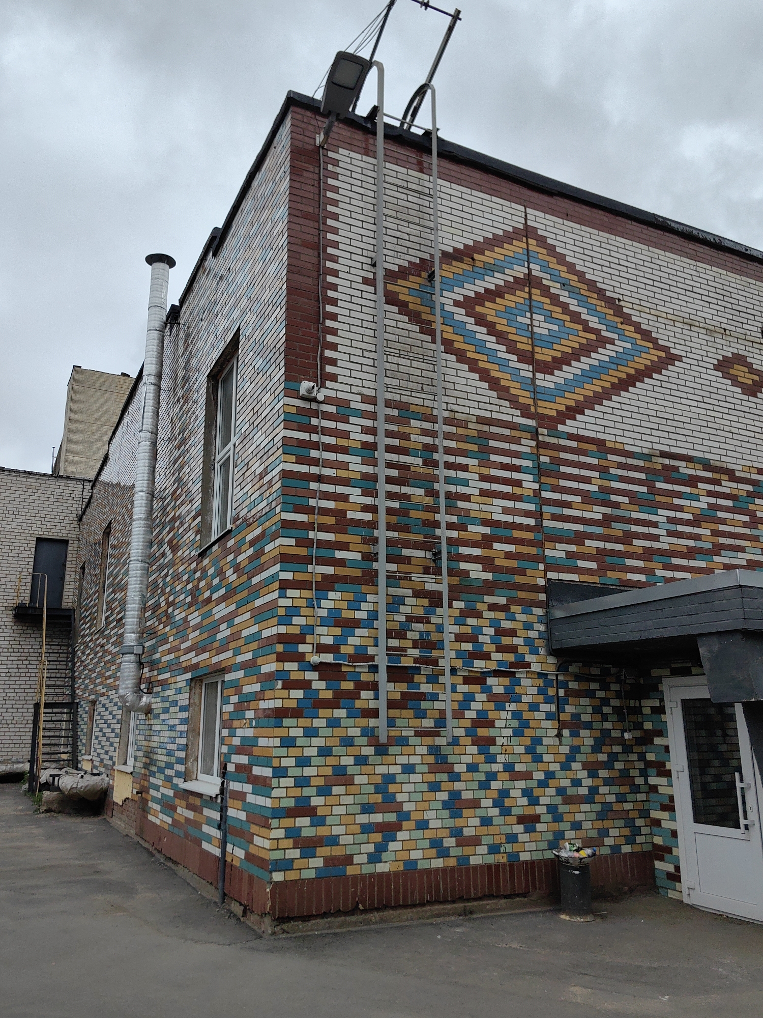The image shows the corner of a brick building in Minsk, Belarus. The building features a multi-colored brick facade with a geometric diamond pattern in shades of red, yellow, blue, and white. A metal ladder is attached to the side of the building, extending to the roof. A ventilation pipe and a security camera are also visible on the building's exterior. The ground in front of the building is paved with asphalt, and a trash can filled with debris is located near a white door on the right side of the image. The sky is overcast and gray.