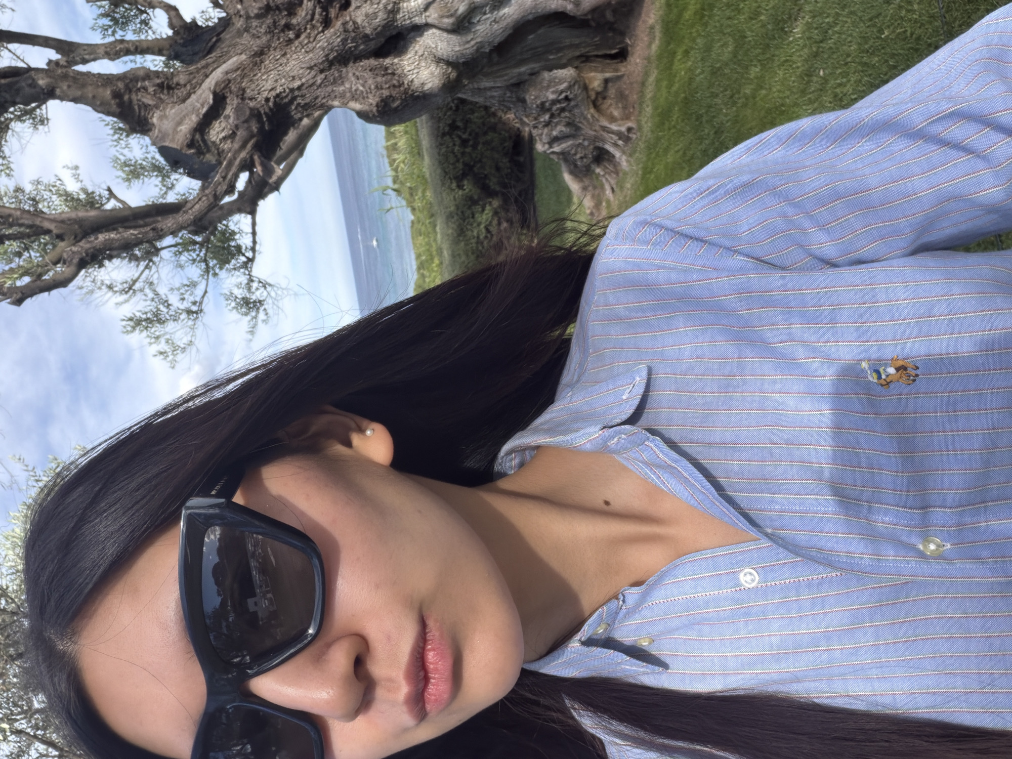 A close-up, slightly angled selfie shows a woman with long, dark hair, wearing dark sunglasses and a light blue striped button-down shirt, possibly cotton, in an outdoor setting. The shirt features a small embroidered logo. Behind the woman, a gnarled tree with thick branches occupies the upper left of the frame, while a glimpse of the ocean and greenery suggests the location is Ramatuelle, France. The sky is partly cloudy. The focus is mainly on the woman's face and clothing.