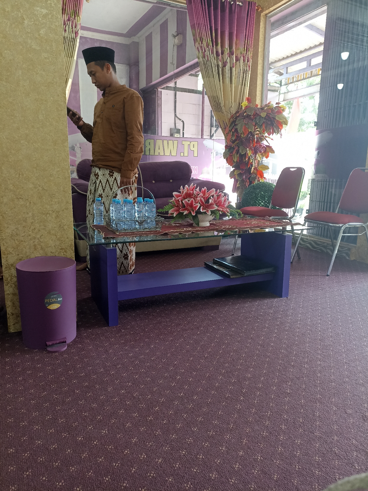 The image depicts an indoor waiting area or reception space, likely within a commercial establishment or office in Tompokersari, Indonesia. A man wearing a brown long-sleeved shirt and a black peci (cap) stands to the left, holding a mobile phone in his left hand. He is positioned near a cream-colored pillar and a purple pedal bin with a "PEDAL Bin" sticker. In the background, a glass-topped coffee table with a purple base displays a vase of pink lilies, several bottles of water in a wire basket, and a dark-covered item. Behind the table, a plush purple sofa is partially visible. The room features striped purple and white walls, and heavy patterned curtains in shades of purple and yellow hang near a large window or glass partition, through which daylight is visible. Two red chairs with chrome legs are situated in the mid-right of the frame. The floor is carpeted in a deep purple with a subtle, repeating pattern. A sign with the text "RAW.TP" is faintly visible on the wall behind the sofa.