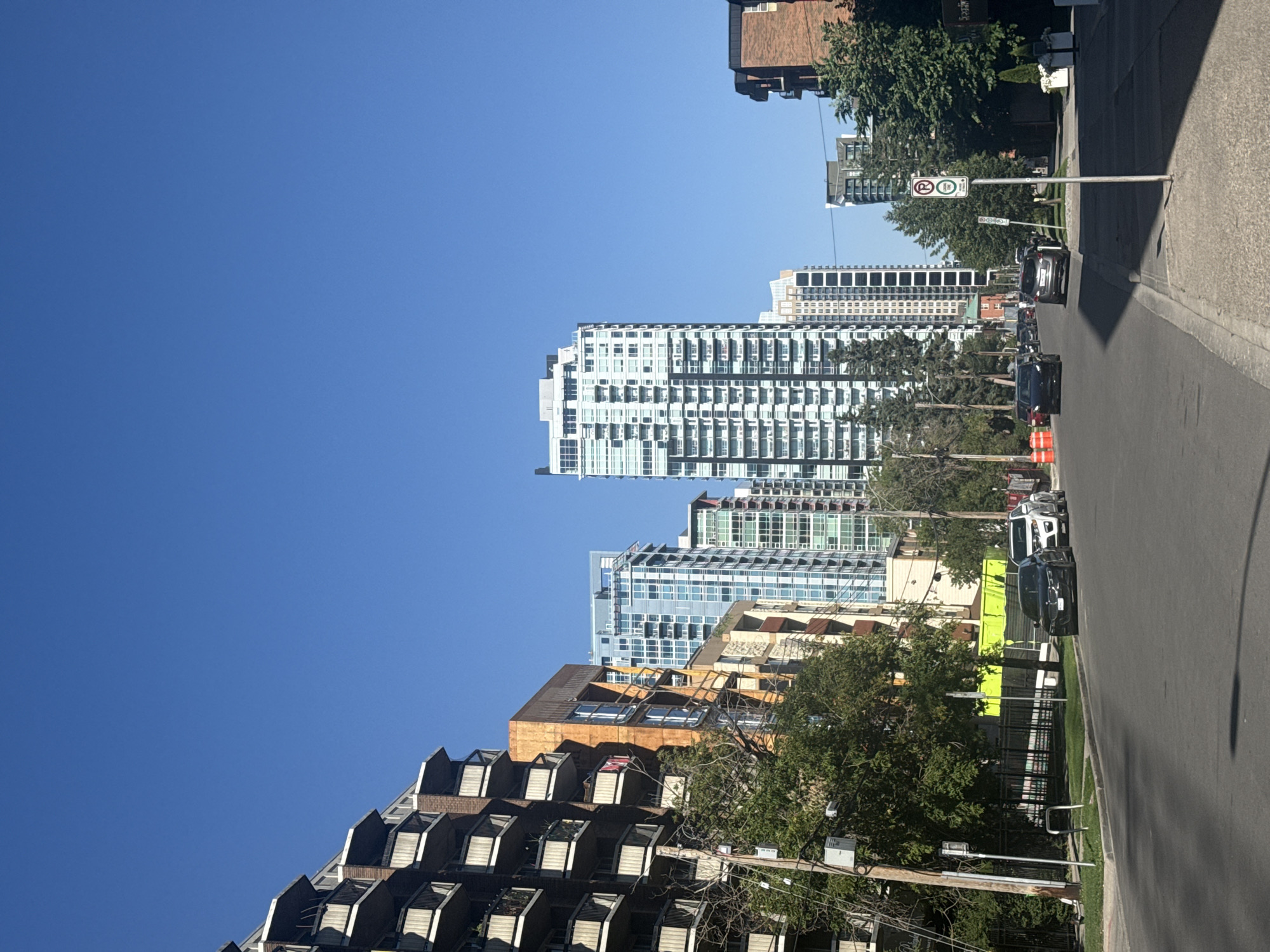 The image depicts a street scene in the Beltline neighborhood of Calgary, Alberta, Canada, on a clear, sunny day. The perspective is from the street, looking down a gentle incline towards a cluster of modern high-rise buildings.
On the left side of the street, a multi-story apartment building with numerous balconies is visible. To its right, a taller, more contemporary building with a distinctive orange facade stands out. Further down the street, several sleek, glass-and-steel high-rise residential towers dominate the skyline, characteristic of the Beltline's urban development.
The street itself is paved, with parked cars lining the right side. A variety of vehicles are present, including a black sedan, a silver SUV, and a white sedan. A street sign indicating parking restrictions (no parking between certain hours) is prominently positioned on the sidewalk on the right.
Trees line the sidewalks, providing greenery against the backdrop of the urban architecture. The sky is a vibrant, cloudless blue. The overall impression is one of a well-maintained, modern urban environment.