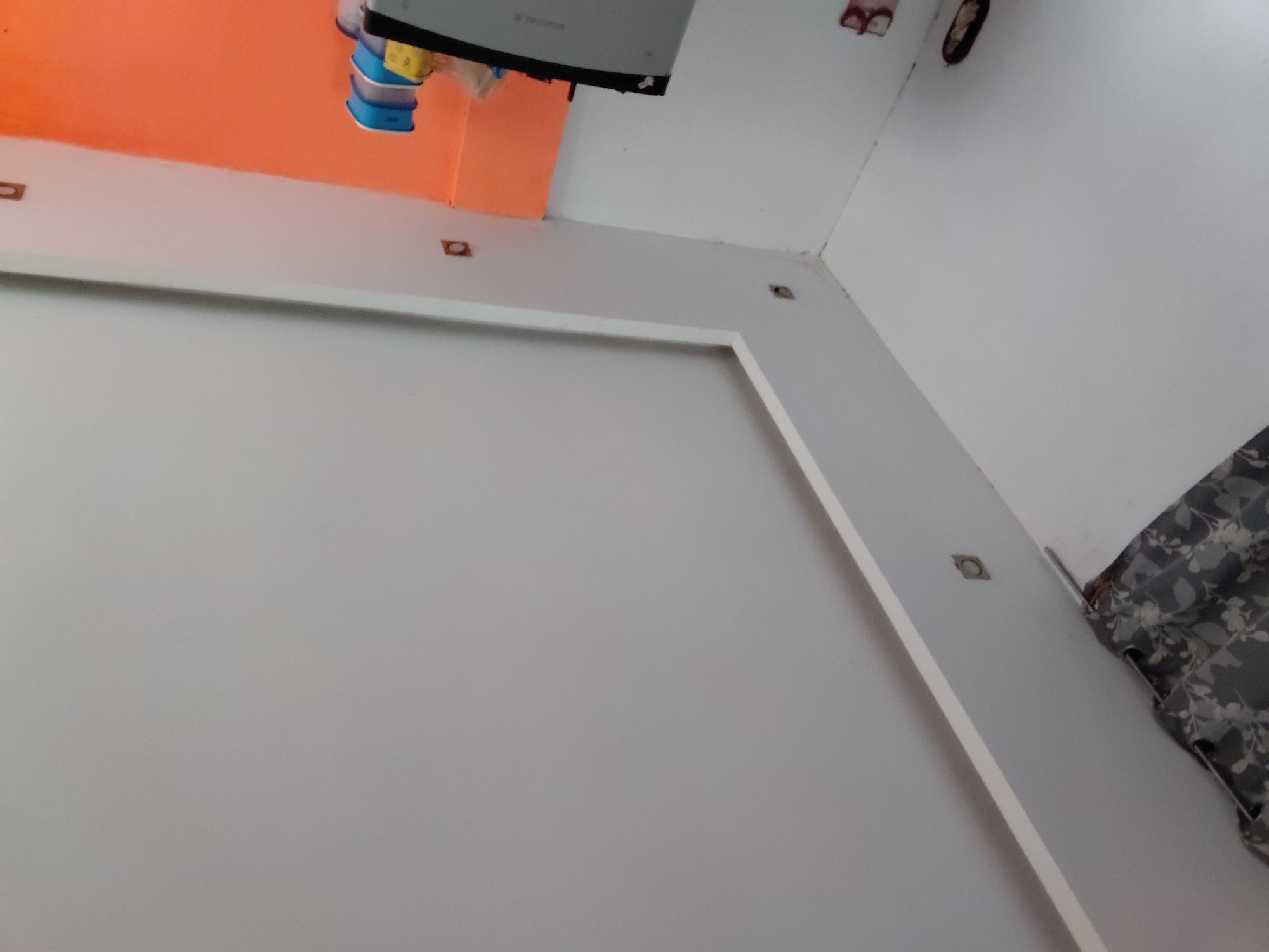 The image captures a tilted, low-angle perspective of an indoor room in Santa Ana, Venezuela. The dominant feature is a large, light grey wall, which includes a rectangular, raised white trim or molding, creating a framed effect. Several small, square recessed light fixtures are visible within this wall and the adjacent white ceiling structure.
In the upper-left area of the image (which would be the top-left corner of the room if correctly oriented), a portion of a brightly orange-painted wall is seen. Above this orange section, an electronic device, appearing to be a Wi-Fi router or similar network equipment, is mounted or hanging. It has distinctive blue and yellow components or cables attached. The text "TECHNOS" and "5G" is faintly visible on its surface.
Further to the upper right, another small, dark recessed fixture is present in the white ceiling. On the far right edge of the frame, a grey curtain with a white floral pattern is partially visible, indicating a window or doorway nearby. No people are present, and no activity is taking place; the image is a static interior shot. The room exhibits a contemporary design with functional elements.