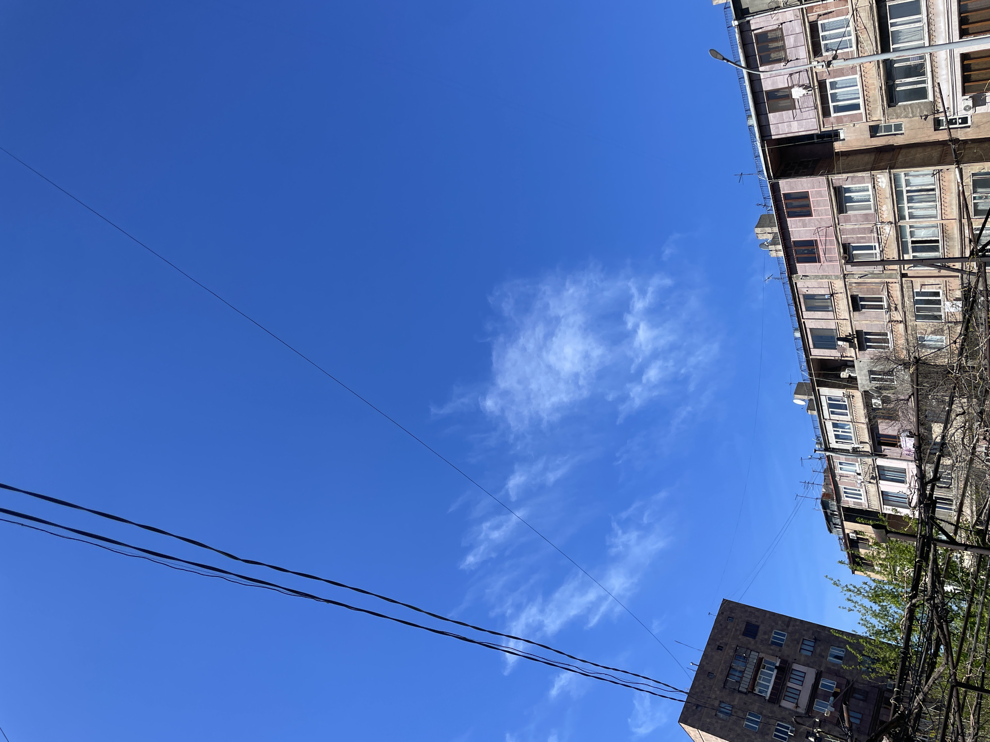 The image captures an upward view of an urban landscape under a vibrant blue sky, adorned with scattered white, wispy clouds.
A tall, multi-story residential building occupies the right side of the frame. It appears aged, featuring numerous windows, some with curtains, and multiple balconies. Air conditioning units are visible on its exterior, and its facade has a faded brownish-pink hue. A street lamp post extends vertically alongside the building.
In the lower left foreground, another, darker-colored building, possibly made of stone or concrete, is visible with a pattern of rectangular windows and some balconies. Green foliage from a tree partially obscures the lower section of this building.
Several dark power lines or communication cables crisscross the sky, running diagonally across the middle and lower portions of the frame. The bright sky and clear visibility indicate the scene takes place during the daytime. No people are visible, and there is no discernible text. The architecture suggests a typical urban residential area, consistent with Yerevan, Armenia.