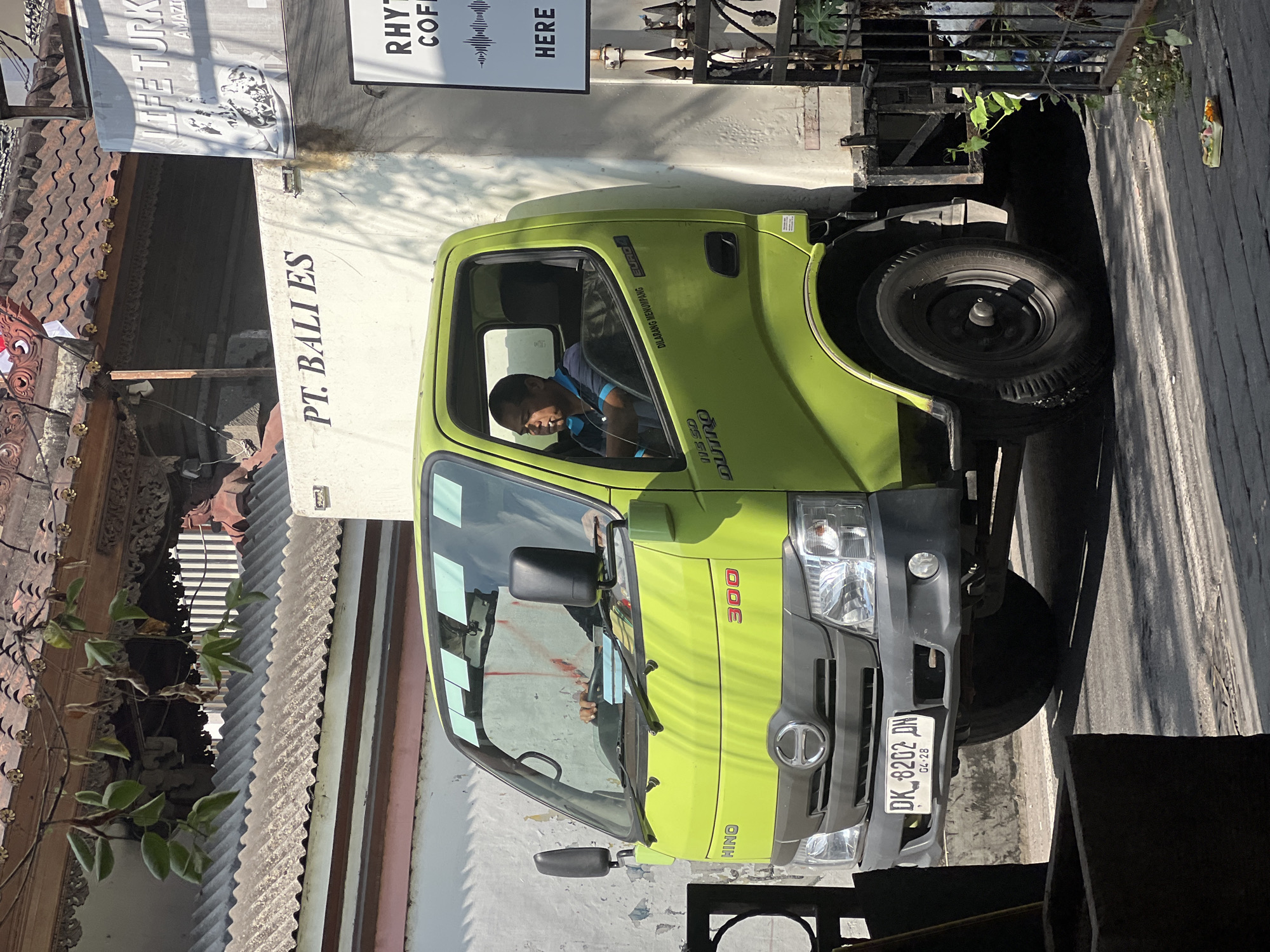 In the city of Kuta, Indonesia, a bright lime green Hino 300 Dutro truck is parked on a street. The truck has a white cargo box mounted on its chassis. The license plate reads "DK 8202 DN". A man is visible inside the driver's cabin, wearing a blue and black patterned short-sleeved shirt and holding a cigarette. The truck's windows are adorned with several square stickers, possibly for toll or parking permits. To the right of the truck, a sign displays the text "RHYTHM COFFEE HERE". In the background, traditional Balinese architecture with red tile roofs and intricate carvings is visible. The scene is illuminated by bright sunlight, casting distinct shadows on the street.