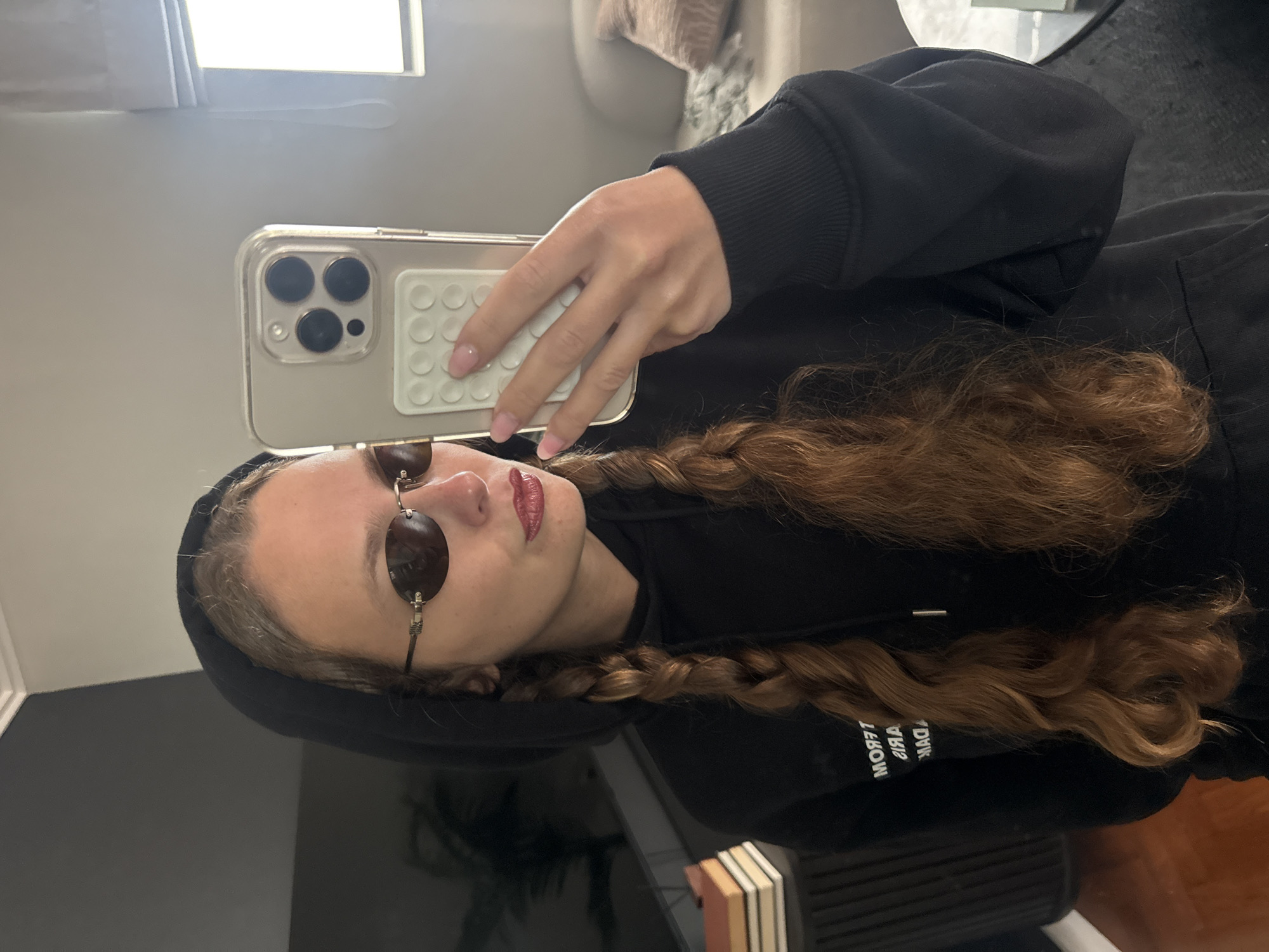 This media file is a mirror selfie taken indoors, featuring a young person, likely a female, in Cape Town, South Africa. The person is holding a light-colored smartphone with a clear case and a white, textured grip on the back, visible in the reflection. They are wearing a black hoodie, small oval-shaped dark sunglasses, and red lipstick. Their long, wavy auburn hair is styled in two braids. Light pink nail polish is visible on their fingers.
The activity taking place is the act of taking a self-portrait in a mirror. The person's face is partially obscured by the sunglasses, making their direct expression unclear, but the overall presentation suggests a casual yet styled look.
Notable details include a bright, rectangular light source reflecting at the top of the mirror. A blurred background behind the person shows what appears to be a light-colored couch or upholstered furniture. Below the person's reflection, a dark, reflective surface, possibly a table, shows stacked items resembling books. Visible text on the black hoodie is reversed due to the mirror, appearing as "MODFT" and "OVIA," likely part of a brand name or phrase. The setting provides no specific outdoor or time-of-day cues beyond being indoors.