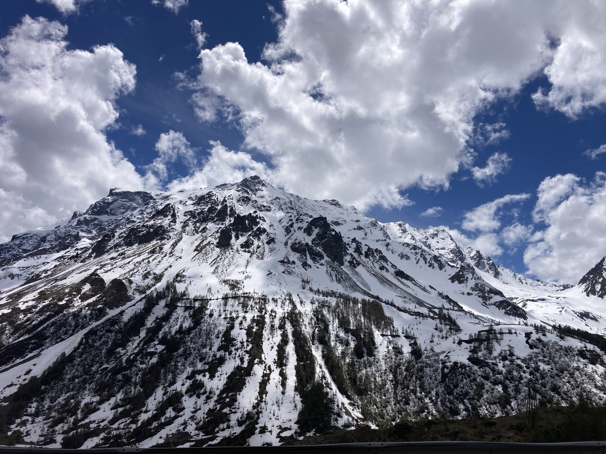 A landscape shot displays a snow-laden mountain range under a sky filled with cumulus clouds. The sky is vibrant blue, interspersed with patches of white fluffy clouds that occupy a significant portion of the frame. The mountains are the primary subject, exhibiting dark rocky patches emerging from the snow cover. Sparse trees dot the mountainside, especially towards the base where snow recedes to reveal the terrain. The photograph is taken from a slightly low angle, enhancing the imposing presence of the mountains. The composition captures the contrast between the bright sky and the snow-covered mountains, emphasizing the natural beauty of the landscape. The location is Unknown, Unknown.