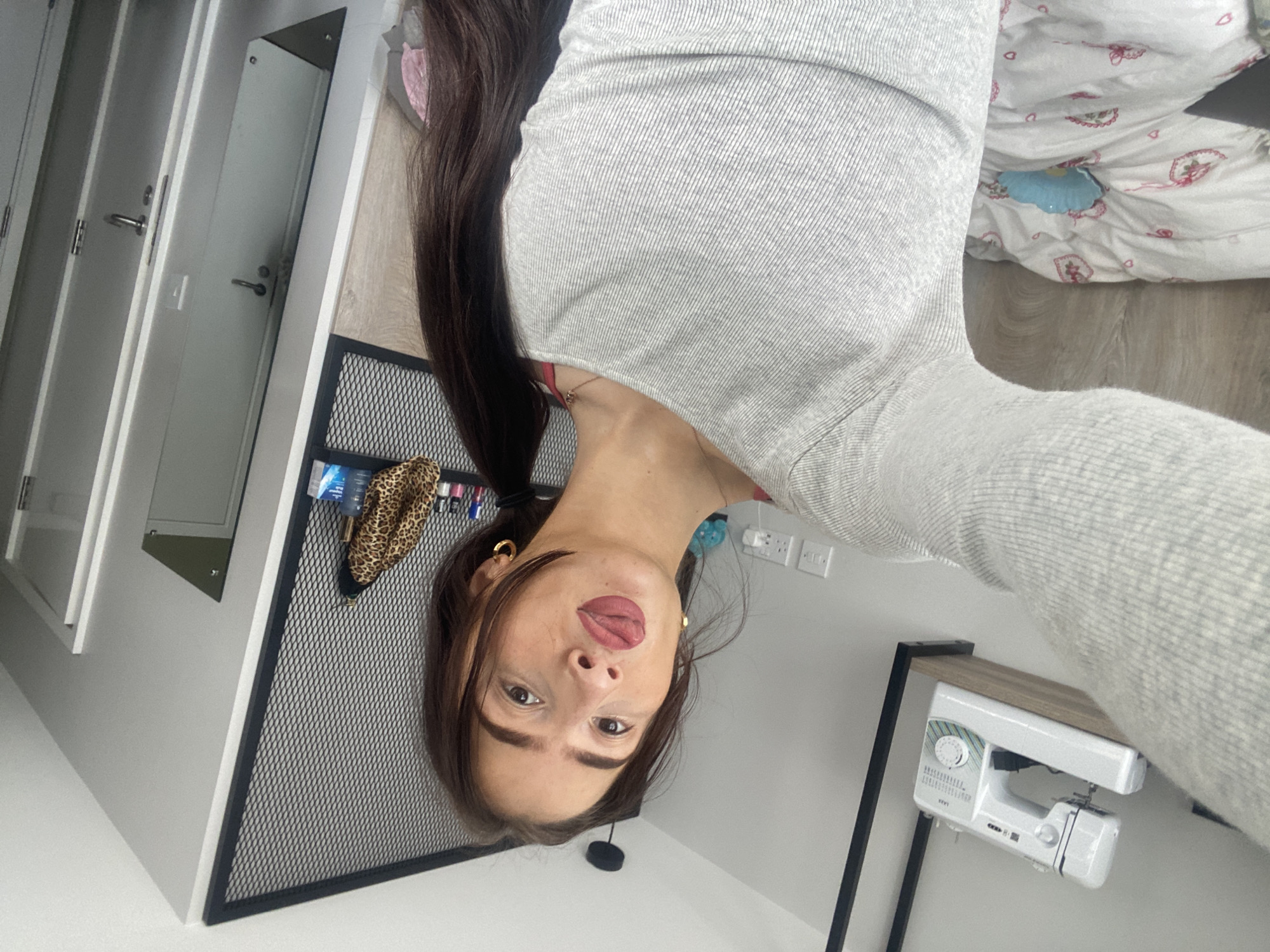 A woman with brown hair is taking a selfie indoors. She is wearing a gray long-sleeved shirt. Behind her, there is a bed with a patterned comforter, a black metal shelf, and a white sewing machine on a wooden table. To the left, there are white doors and a mirror. The location appears to be a residential space, but there are no indicators as to the location of this room, including city or borough of London, United Kingdom.