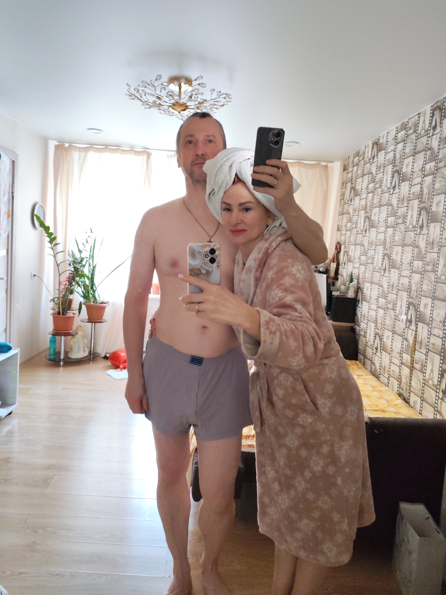 A selfie, taken indoors, features a middle-aged man and woman. The man, fair-skinned, is shirtless except for gray boxer shorts. The woman, fair-skinned, wears a floral-patterned robe and a white towel wrapped around her head. She holds up two smartphones to take the photo. The background includes a window with sheer curtains, potted plants on a stand, a patterned wallpaper with images, and a ceiling light fixture. The location is Chaikovsky, Russia.