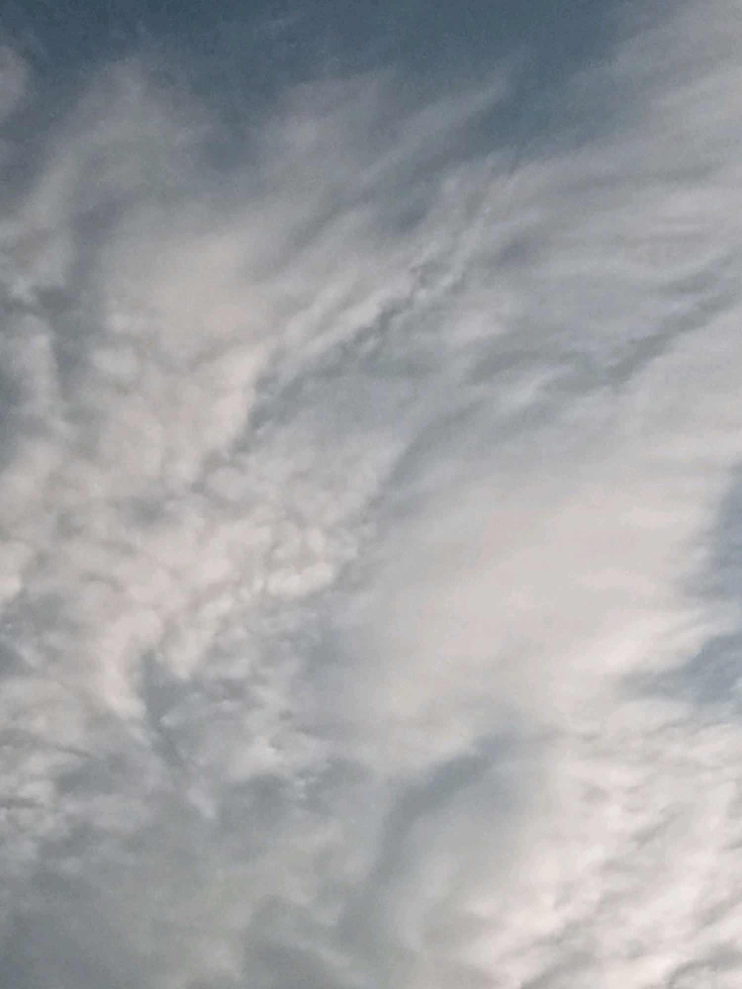 The image is a low-resolution, abstract depiction of a cloudy sky. The top portion of the image shows a darker, deeper blue sky, transitioning into lighter shades of grey and white as it moves downwards. Wispy, scattered clouds dominate the frame, with some appearing denser and more opaque than others. There are no discernible objects, people, or actions visible, and the scene is entirely abstract, suggesting a focus on atmospheric conditions. There is no information within the image that can be used to determine its location in Tando Jam, Pakistan.