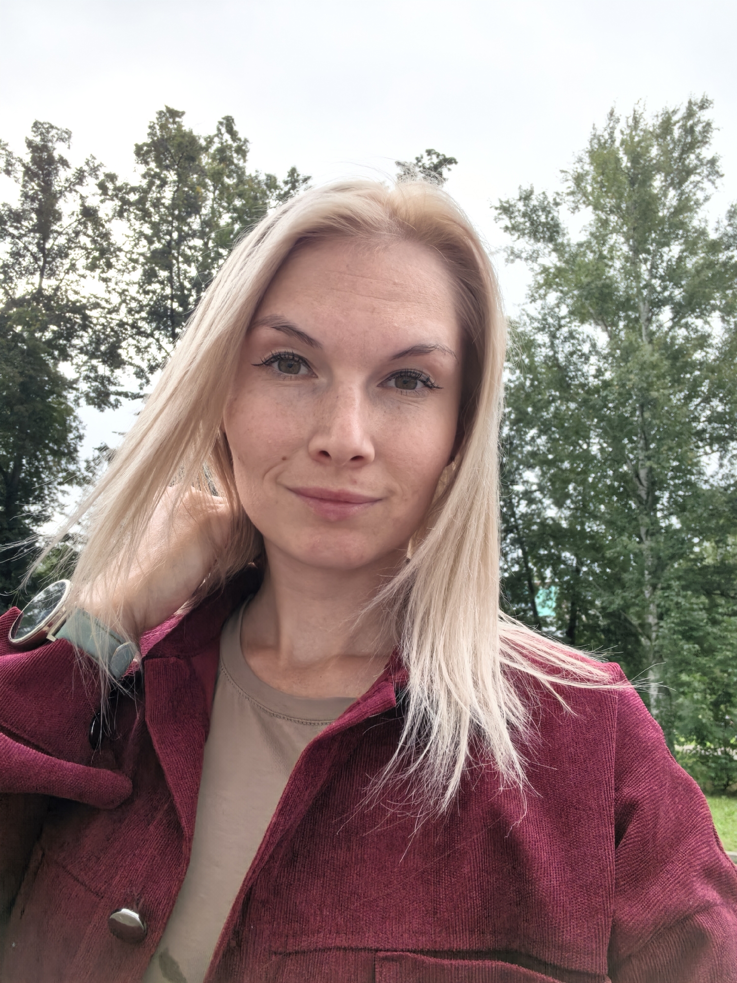 A young Caucasian woman with blonde hair and fair skin, wearing a burgundy corduroy jacket over a tan t-shirt, takes a selfie outdoors. She has light green eyes, subtle makeup, and a light dusting of freckles on her cheeks and nose. Her left hand is raised, with fingers touching her hair. A silver watch with a light blue band is visible on her left wrist. The background consists of green trees and an overcast sky, suggesting an outdoor setting. The image was captured in the city of Sterlitamak, Russia.