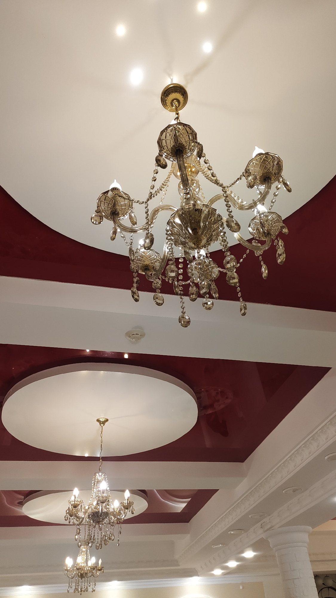 This image shows an interior view of a ceiling in Beryozovka, Russia. The ceiling is decorated with a combination of white and dark red panels, featuring recessed lighting and ornate molding. Two crystal chandeliers are visible, one larger and more elaborate than the other. The chandeliers are suspended from the ceiling and adorned with numerous sparkling crystals. The overall aesthetic suggests a well-decorated and possibly formal interior space, such as a restaurant or banquet hall. The image is taken from a low angle, looking upwards at the ceiling and the chandeliers.