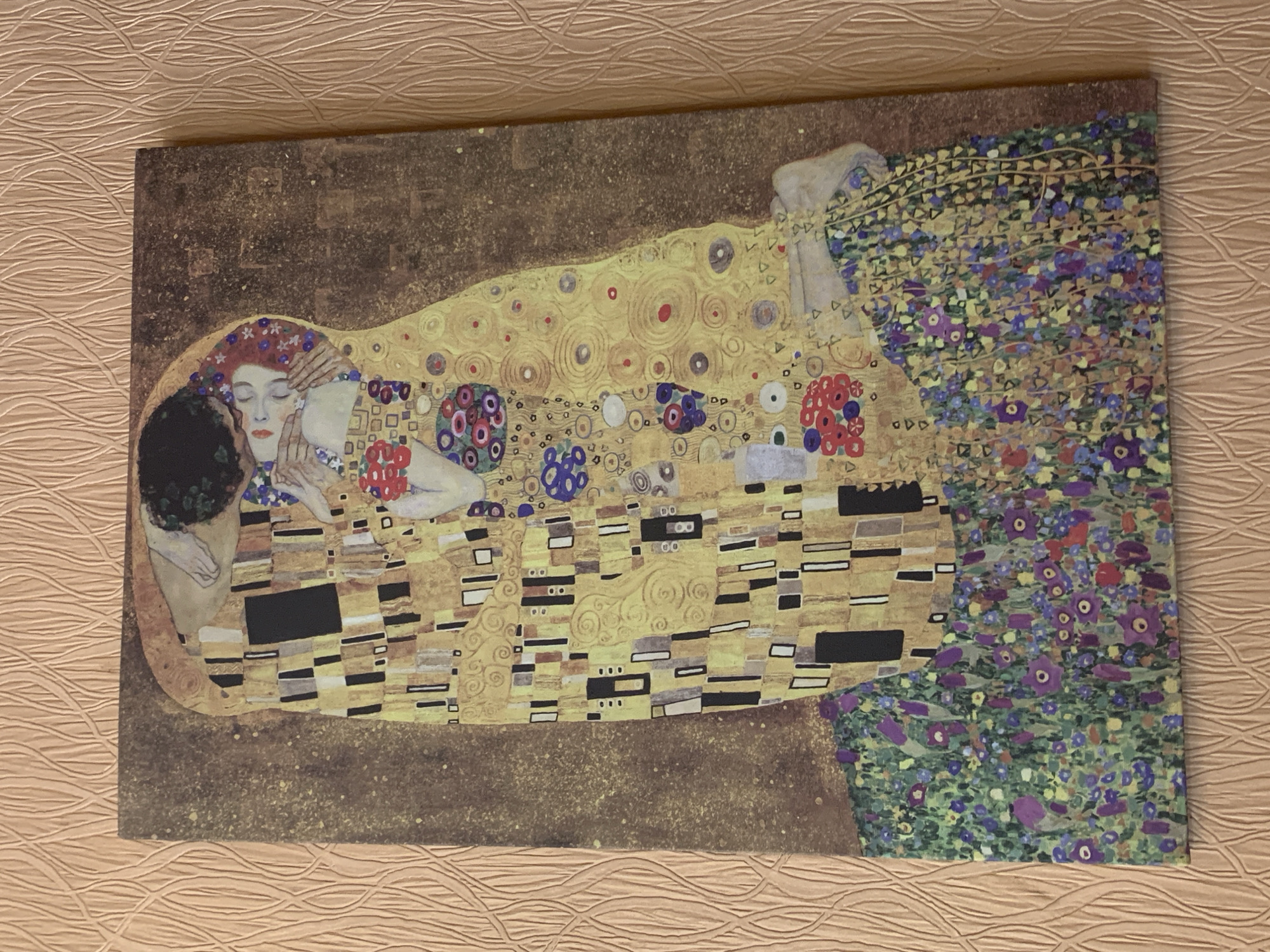 A framed reproduction of Gustav Klimt's "The Kiss" is displayed on a textured, peach-colored wall. The artwork depicts a man and woman embracing, both adorned in elaborate, gold-patterned robes. The man's head is bowed, kissing the woman's cheek, while she appears to be in a state of ecstasy, her eyes closed. They are positioned on a floral carpet, with a dark, speckled background behind them. The frame is not visible. The overall lighting suggests an indoor setting.