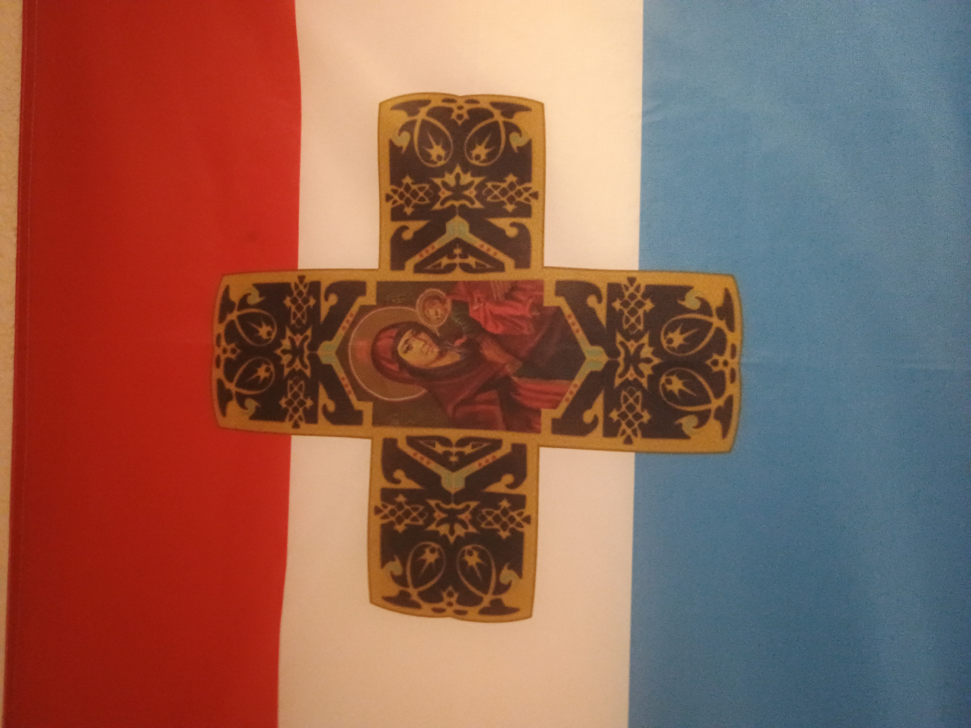 Here is a description of the image:
The image shows a flag with a vertical tricolor of red, white, and blue. Centered on the white band is a decorative cross. The cross has a central panel depicting an icon of the Virgin Mary holding the infant Jesus. The cross also has an intricate, floral-like pattern. The flag is slightly wrinkled and appears to be hanging on a wall.
I have no information about the location of the image.