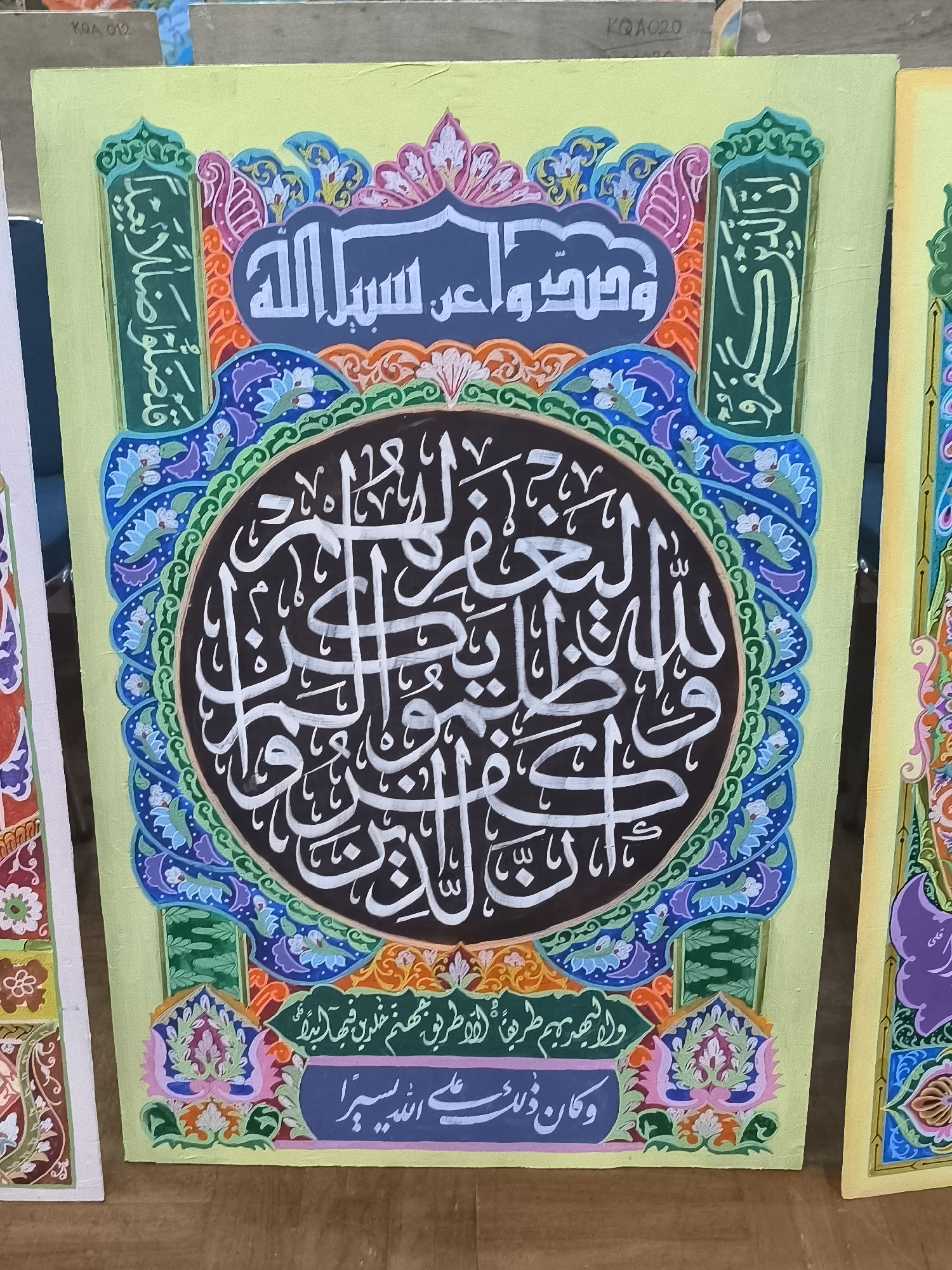 This is a close-up, eye-level, interior shot of a colorful Islamic calligraphy artwork. The artwork features a central oval inscribed with white Arabic script on a black background, surrounded by a vibrant, multi-layered border of floral and geometric patterns in green, blue, purple, orange, and pink. The artwork is displayed on a light yellow canvas. Above the central oval, in a dark blue cloud-like shape, are two lines of white Arabic calligraphy. On the left and right sides of the canvas, flanking the central design, are vertical bands of green with white Arabic calligraphy. At the bottom of the canvas, below the central design, is a dark blue rectangular area containing white Arabic calligraphy. Two other artworks, one on the left and one on the right, are partially visible, suggesting this is part of a collection or exhibition. The background appears to be a wall with some light wooden elements and potentially other canvases. The image does not provide any information about the city of Minasa Te'ne, Indonesia, or any people.