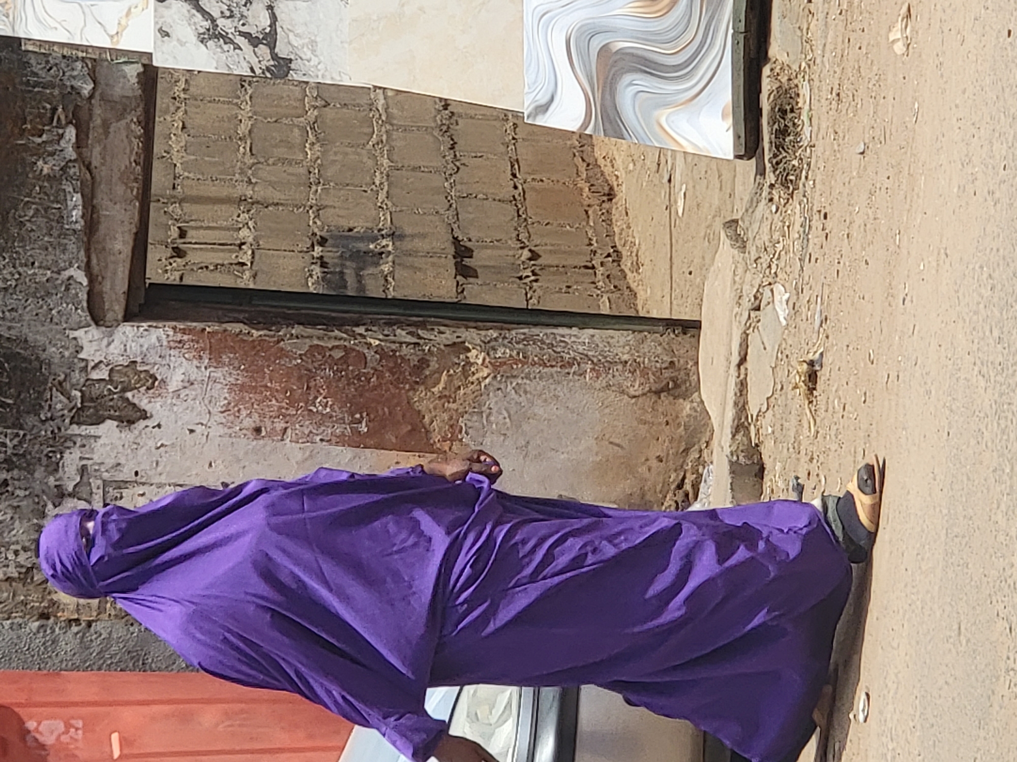 A person clad in a flowing purple garment with a head covering stands on a paved surface, likely a sidewalk or street, in Maiburiji, Nigeria. Their right foot, visible, is shod in a sandal. The background includes a building with a distressed facade, exhibiting exposed brickwork and peeling paint. Ceramic tiles are leaning against the wall of the building. A portion of a motor vehicle is visible towards the bottom of the frame. The environment appears arid and sunlit.