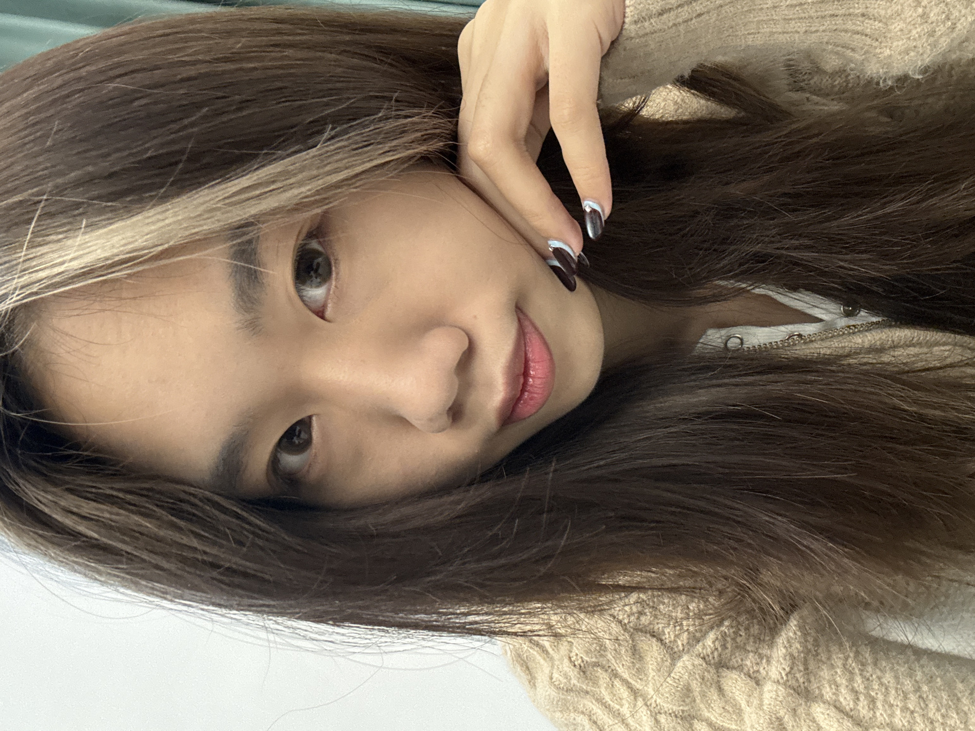 A medium close-up portrait depicts a young East Asian female in an indoor setting. Her long, dark brown hair features a distinct blonde streak on her left side and is parted on her right, falling over her shoulders. Her dark eyes are directed at the viewer, and her lips are tinted pink. She wears a light beige cable-knit sweater over a white undergarment, revealing a silver-colored chain necklace. Her left hand is positioned with fingers resting along her jawline. Her fingernails are manicured with dark polish, accentuated by light blue crescent shapes and small silver details near the tips. The background consists of an out-of-focus plain white surface to her right and a muted light green-blue surface to her left.