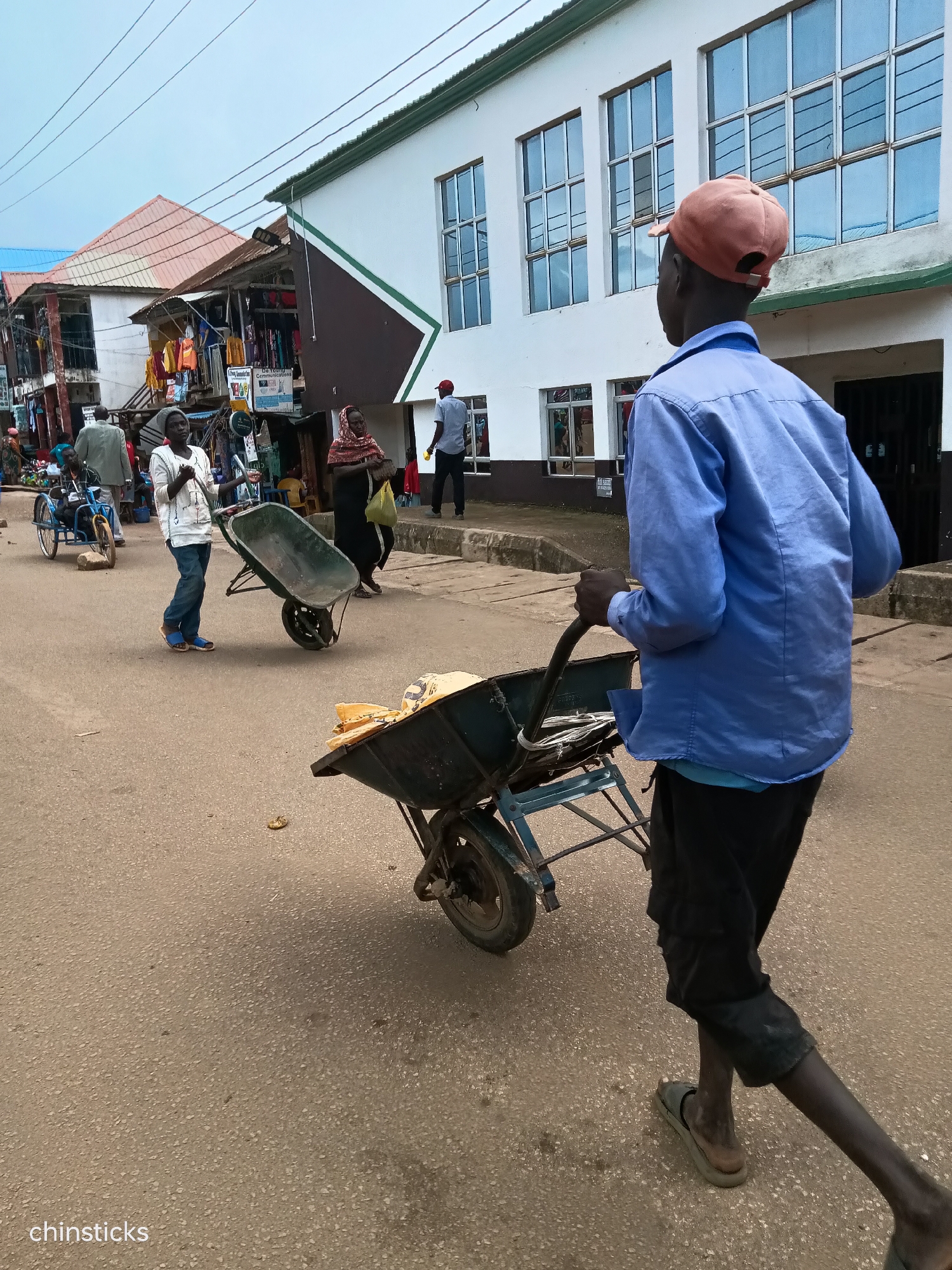 In Bukuru, Nigeria, a man wearing a blue long-sleeved shirt and black shorts pushes a wheelbarrow filled with yellow bags. He is wearing a peach-colored baseball cap and dark sandals. The wheelbarrow has a blue frame and a metal basin. In the background, a white building with large windows, a market with stalls displaying clothing, and other people are visible. The ground is paved with asphalt.