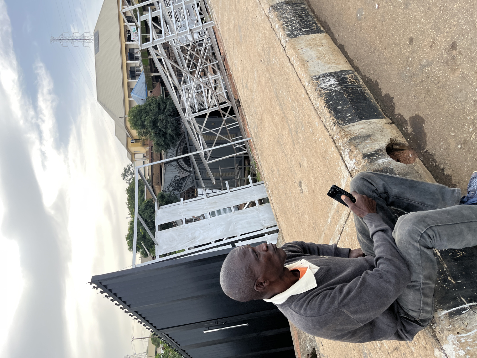 A man sits on a raised concrete curb, looking up and to his right. He is wearing a dark gray sweater over an orange collared shirt, and dark jeans. He holds a black smartphone in his right hand. The man's skin is dark, and he is balding. He appears to be in a relaxed or contemplative pose.
Behind the man, a large, dark structure, possibly a shipping container or a covered stall, is visible. Beyond that, a white metal scaffolding or construction project is partially visible, with trees and buildings in the background. A high-voltage power line tower is prominent in the distance, suggesting an urban or semi-urban environment. The sky is overcast with some white clouds.
The scene appears to be outdoors, likely during the daytime given the lighting conditions. The curb he is sitting on is wide and has alternating black and white painted stripes, with visible wear and tear. The ground surface is rough concrete. The overall impression is of an everyday scene, possibly in a public area where construction or renovation is taking place. No text is clearly visible in the image. The location context provided is Gura Top, Nigeria.