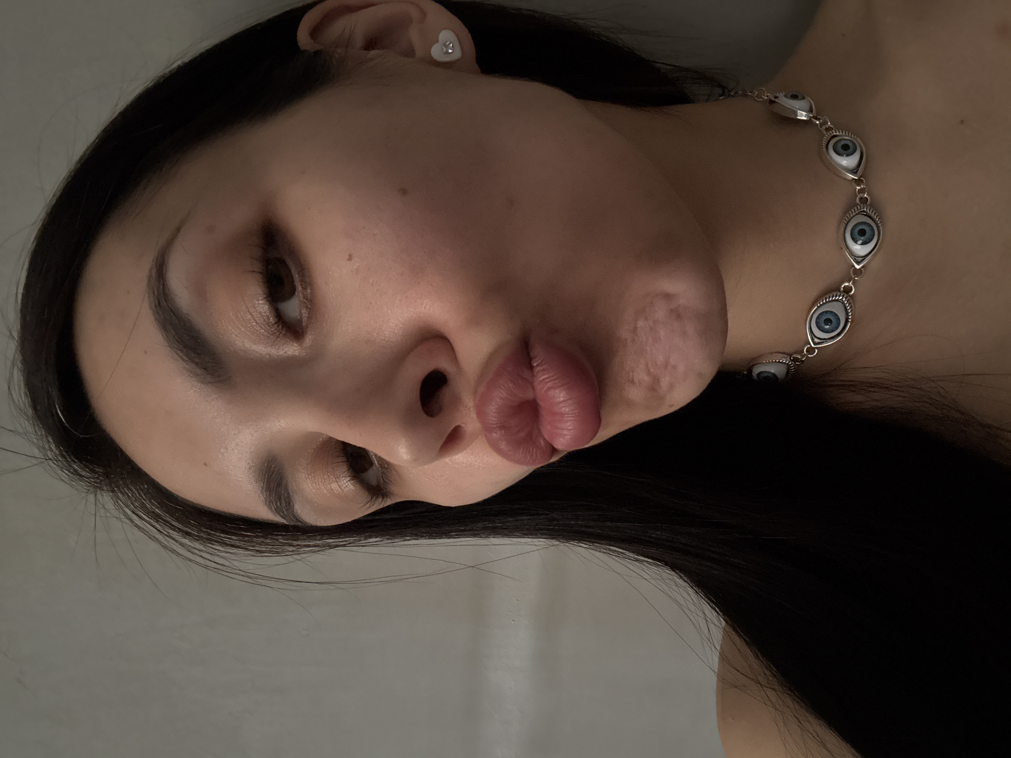 A selfie captures a woman with long, straight, dark hair. She has fair skin, dark eyebrows, and brown eyes. Her lips are pursed in a kissing pose. She is wearing a silver necklace adorned with eye-shaped charms and a small, white heart-shaped earring. The background is a blurred, light-colored wall. There is no information available to associate this image with the city of Votkinsk, Russia.