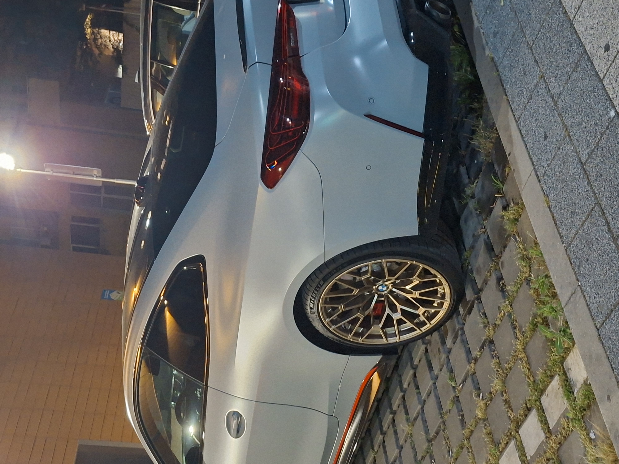 A light gray BMW M4 coupe is parked on a paver stone surface in what appears to be an urban setting at night. The car is positioned at an angle, facing towards the left of the frame, with its rear passenger side prominently displayed. The vehicle has a sleek design with sharp lines, a tinted roof, and distinctive red taillights. Its golden multi-spoke wheels with red brake calipers are visible. An orange accent stripe runs along the lower side of the car.
The environment includes a brick wall in the background to the left, a lamppost with a light illuminating the scene, and another car partially visible behind the BMW. The ground is covered in square gray pavers with patches of grass growing between them. A gray concrete curb and sidewalk are in the foreground. The overall impression is one of a street or parking area at dusk or after dark, with artificial lighting casting shadows and highlighting the car. The location is identified as Plovdiv, Bulgaria.