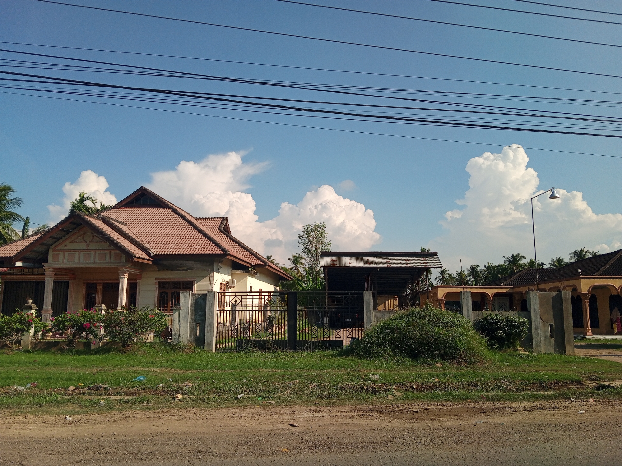 The image depicts a street scene in Lhoknga, Indonesia, on a clear day with a blue sky dotted with white, fluffy clouds. In the foreground, a dirt road with some scattered debris runs horizontally across the bottom of the frame. Beyond the road is a strip of green grass, also with some litter.
The middle ground features several structures. On the left is a beige house with a reddish-brown tiled roof, featuring a covered porch supported by pillars. A black metal fence encloses the property, with lush green bushes and flowering plants visible behind it. To the right of this house is an open-sided structure with a corrugated metal roof, likely a carport or workshop, where a dark-colored vehicle is parked. Another building, possibly a mosque or community hall, is visible further to the right, characterized by its long, covered arched walkways and a similar reddish-brown tiled roof. A street lamp stands tall in front of one of the buildings. Palm trees are scattered throughout the background, suggesting a tropical or subtropical environment. Several power lines stretch across the top of the image, intersecting the sky. No people are clearly visible or identifiable in the scene. The overall impression is one of a residential or community area in a coastal town in Indonesia.