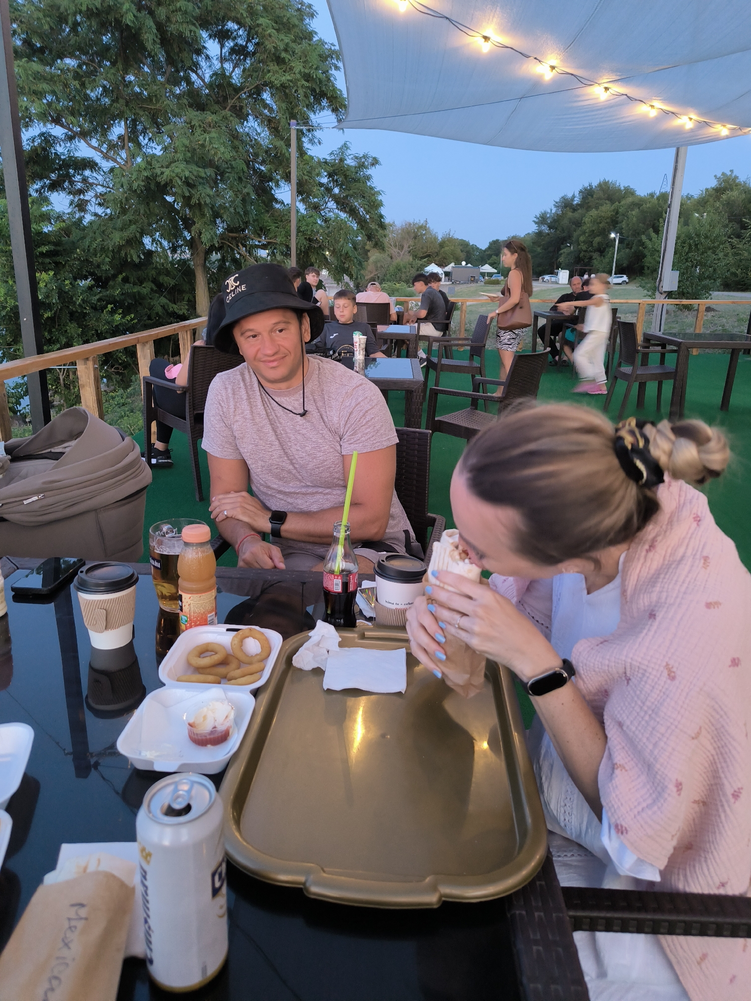 In the city of Ustia, Moldova, at an outdoor dining establishment, two individuals are seated at a table. The man, wearing a black hat and a light pink t-shirt, is looking at the camera. In front of him are a glass of beer, a bottle of juice, onion rings, and a cup. A woman, in the foreground with her back to the camera, is wearing a pink garment and is eating. In front of her is a takeaway food item. The table also holds a can of soda and a coffee cup. The setting appears to be an open-air venue with tables and chairs, a railing, and trees in the background, under a white canopy with string lights.