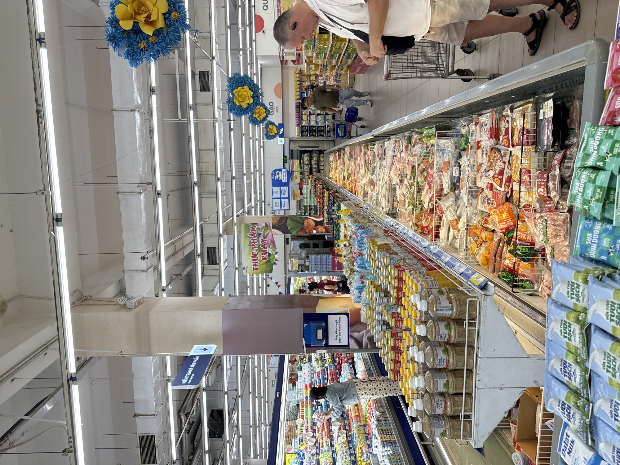 This is a medium-wide shot of the interior of a brightly lit supermarket in Nha Trang, Vietnam, likely a Co.opmart based on the signage. The perspective is from a slightly elevated, eye-level view, looking down a long aisle of refrigerated display cases filled with packaged food items, predominantly meats and prepared meals.
In the foreground, a man wearing a white t-shirt, light-colored shorts, and black sandals is seen from the waist down, facing away from the camera. He is holding a black bag across his chest and is interacting with the contents of a shopping cart. To his left, a row of bottled sauces, labeled "KEWPIE" and in Vietnamese, are prominently displayed. Below these, stacked packages of what appear to be milk cartons, with Vietnamese text and images of cows, are visible.
Further into the aisle, other shoppers are present. A woman in a denim jacket and patterned pants is standing in front of a display of dairy products on the left side of the image. Another person, wearing a dark shirt, is pushing a shopping cart in the background, walking towards the rear of the store. The aisles are lined with extensive refrigerated units displaying a variety of packaged food, including what appears to be fresh meat, seafood, and processed food items.
Overhead, fluorescent lighting illuminates the space. Decorative floral arrangements, one predominantly blue with yellow accents, hang from the ceiling. Signage is visible throughout the store, with some in Vietnamese indicating product categories or store information. One sign in the mid-ground reads "THUC PHAM CHAY," meaning "vegetarian food." The overall environment is typical of a modern, well-stocked supermarket.