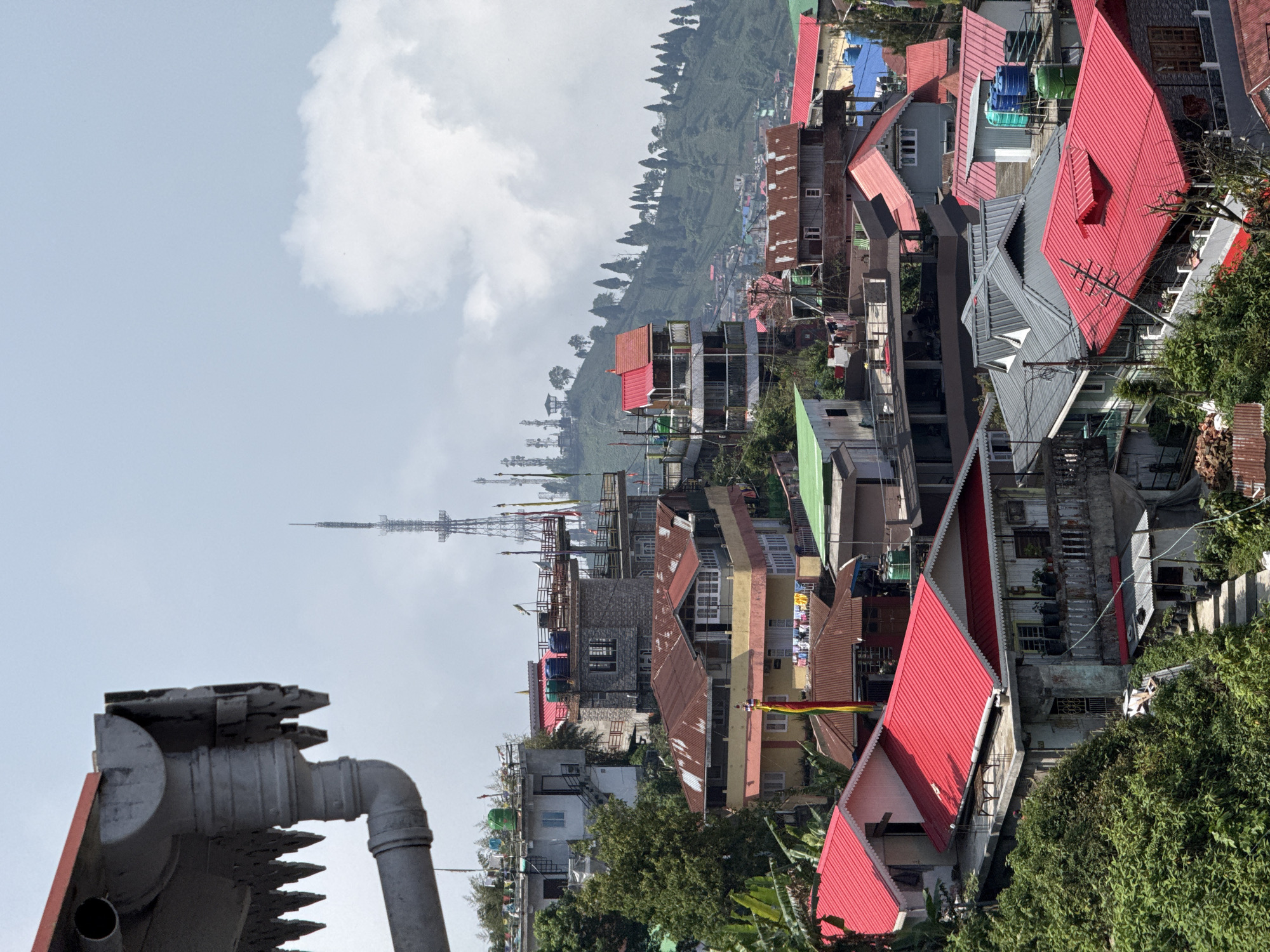 This image captures a panoramic view of a densely populated hillside town, likely Kurseong, India. The scene is presented during daytime under a partly cloudy sky.
In the foreground, to the left, a large, grey, industrial-looking metallic structure with pipes and jagged elements partially obstructs the view. The midground is dominated by numerous multi-story buildings, clustered tightly together on the sloping terrain. These houses feature a variety of architectural styles, with many distinguished by their sloped roofs, predominantly in vibrant red, but also in grey, green, and weathered brown. Blue and green water tanks are visible on several rooftops, suggesting residential use. Clotheslines with drying laundry can be seen on some balconies and roofs. A tall, slender communication or broadcast tower, constructed from a lattice framework, stands prominently in the center-left of the townscape, extending upwards into the cloudy sky.
In the background, steep, verdant hillsides rise, covered in dense foliage and distinct rows of tall, slender trees, possibly indicative of tea gardens or forests common to the region. The sky above is a light blue-grey, dotted with fluffy white clouds.
A notable detail among the buildings is a colorful banner displaying horizontal stripes of red, yellow, and green, which could be a decorative flag or a prayer flag. No individuals are visible in the image. While some indistinct markings appear on distant buildings, no clear, readable text is discernible.