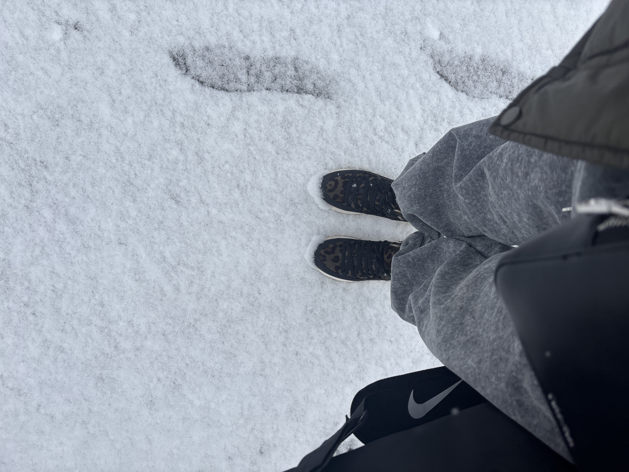 The photograph shows a first-person perspective of a person standing in snow. The person is wearing gray sweatpants and black sneakers with a brown animal-like print. A black Nike bag hangs from the person's shoulder. Footprints are visible in the snow. The location is likely Perm, Russia.