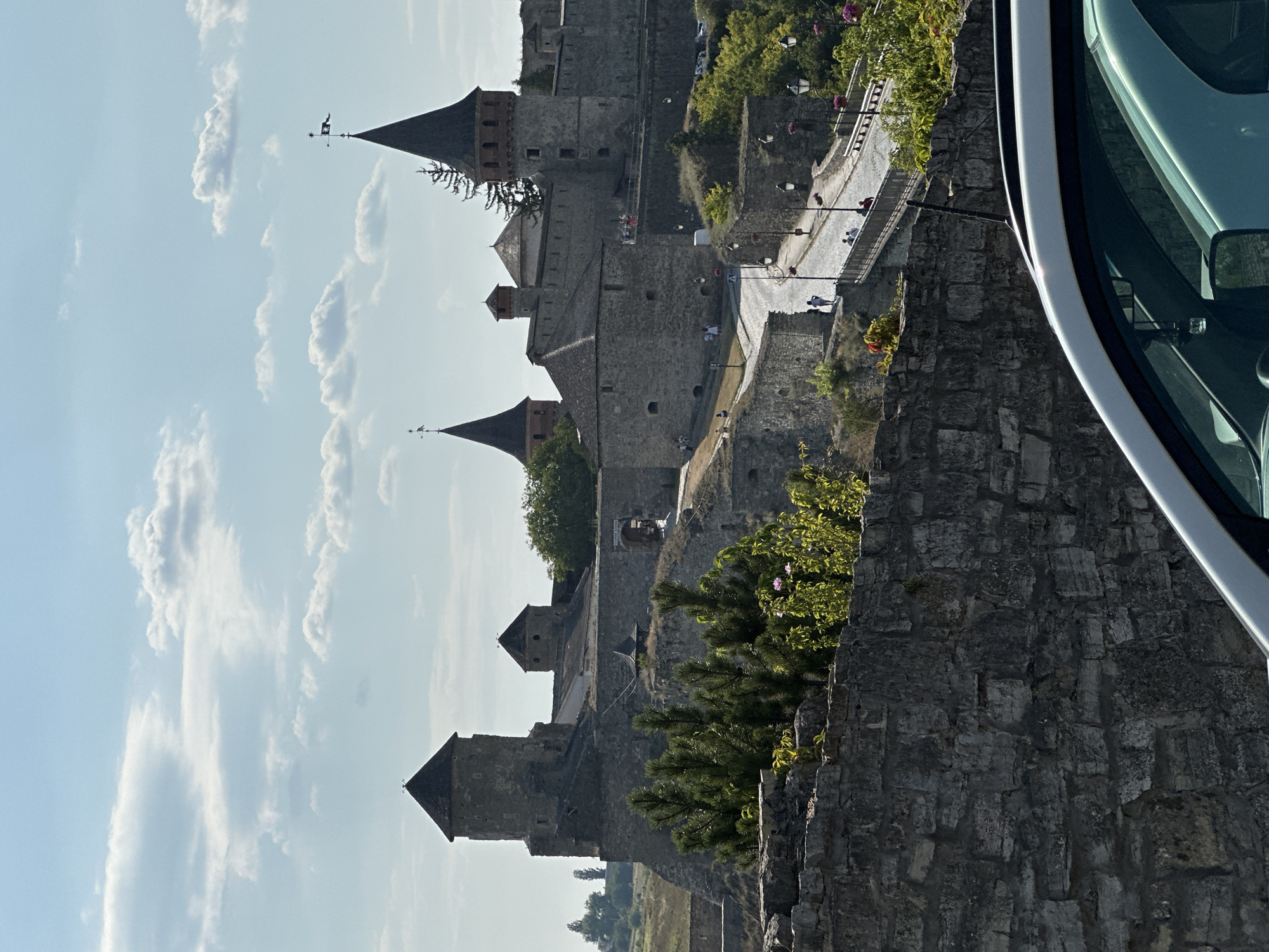 This is a vertical, eye-level, full-shot photograph of the Kamianets-Podilskyi Castle in Ukraine on a bright, cloudy day. The foreground is dominated by the hood and windshield of a white vehicle, obscuring the bottom left of the frame. Beyond the vehicle is a low, rough stone wall that runs horizontally across the lower portion of the image.
Behind the wall, the castle complex is visible, perched on a rocky incline. It is constructed of grey stone and features multiple towers with dark, conical roofs. The tallest tower is on the left, and to its right are smaller buildings and another prominent tower with a flag atop it. A paved road winds its way up to the castle entrance, which is framed by an archway. Several people are walking on the road and the bridge leading to the castle, suggesting it is open to visitors.
The landscape surrounding the castle is hilly and covered in green vegetation, with trees interspersed throughout. The sky is a pale blue, with scattered white, fluffy clouds. The sunlight casts shadows, indicating a mid-day or afternoon setting. The overall impression is one of historical grandeur and a popular tourist destination.