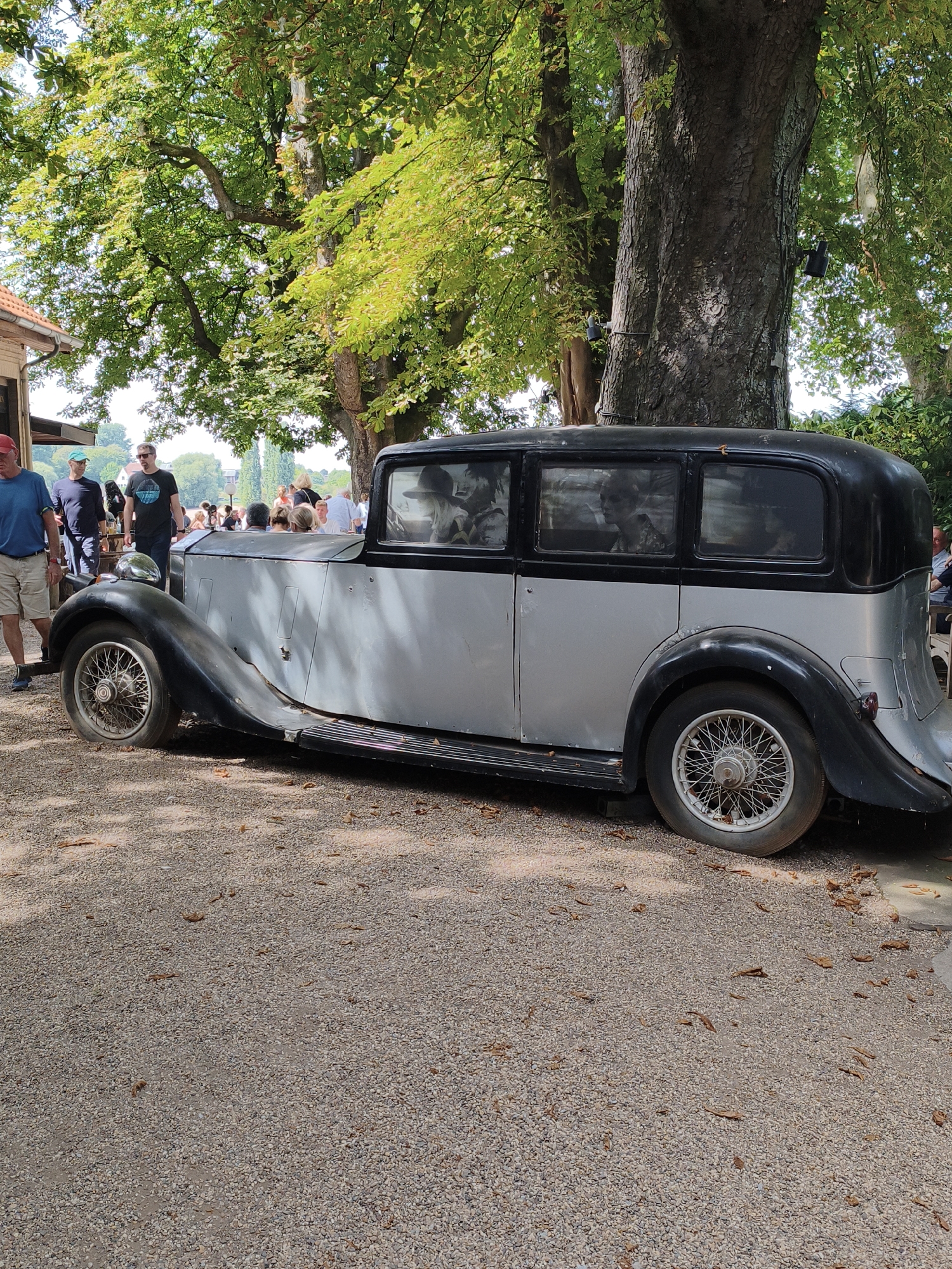 In a sun-dappled outdoor setting, a vintage, two-tone black and silver automobile is parked on a gravel surface. The car, likely a sedan with multiple windows, appears to be from the early to mid-20th century, characterized by its large, rounded fenders and spoked wheels. Several people are visible in the background, some standing and others seated at tables, suggesting a social gathering or event. The environment is lush with mature trees providing ample shade, and foliage is visible throughout the frame. The building on the left suggests a possible cafe or restaurant. There is no explicit indication of the city of Dusseldorf, Germany, in the image itself.