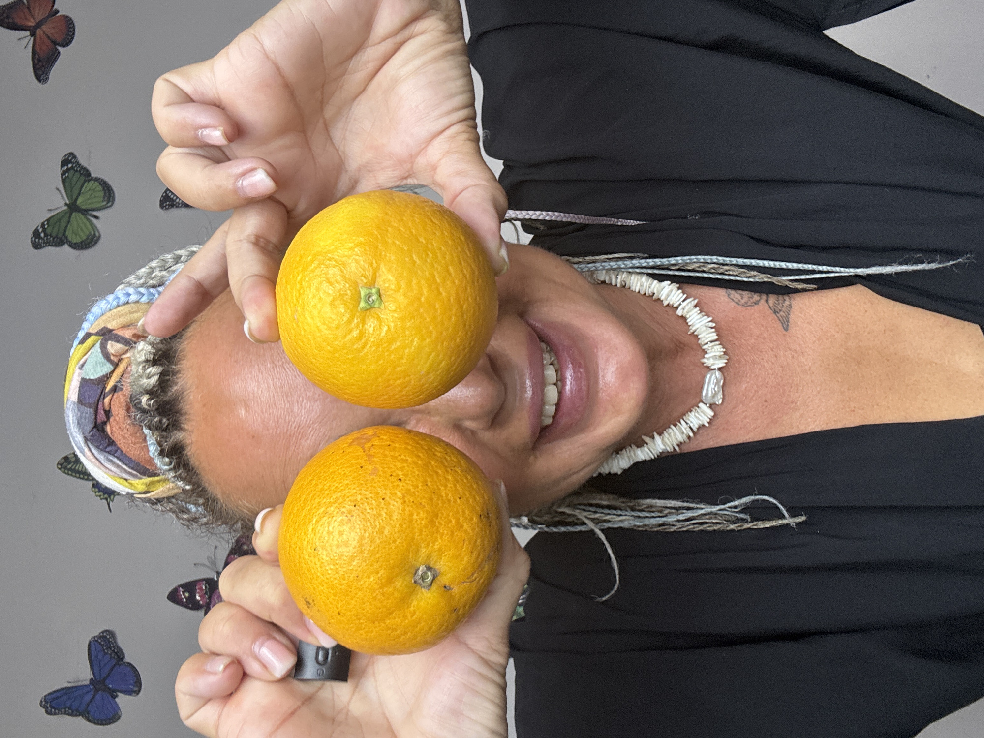 A woman with braided hair and a colorful headscarf is holding two oranges up to her eyes. She is wearing a black top and a white seashell necklace. The background is a gray wall with several butterfly decals. The image is a close-up shot, and it is not possible to determine the specific location within Kuta, Indonesia.