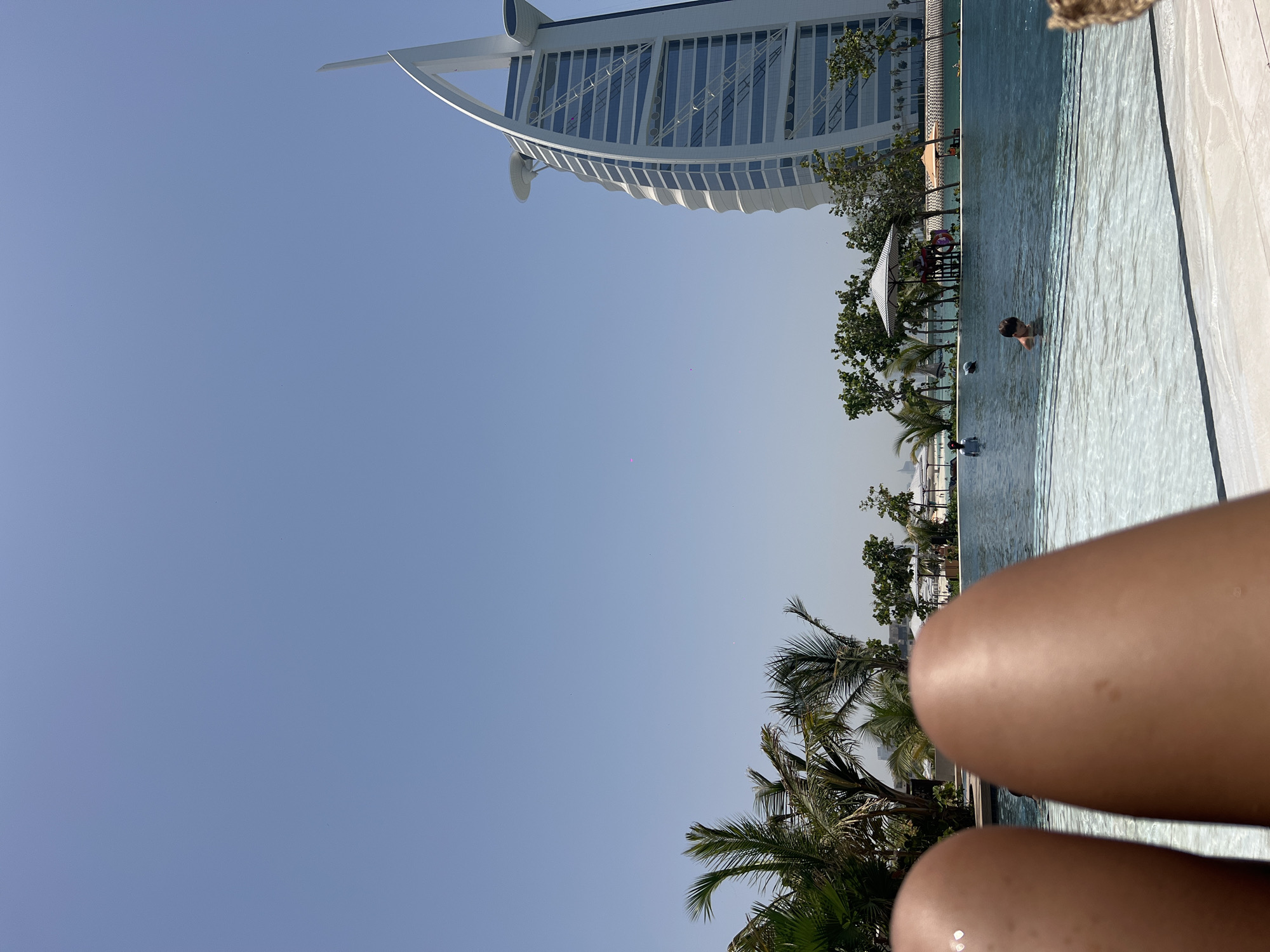 Here's a detailed description of the image:
The image is an eye-level shot taken from a poolside location. A person is seen swimming in the pool. In the background, the Burj Al Arab hotel is clearly visible, placing the location in Dubai, United Arab Emirates. Palm trees and other greenery surround the pool. In the foreground, the legs of the person taking the photo are visible. The sky is a clear, pale blue.