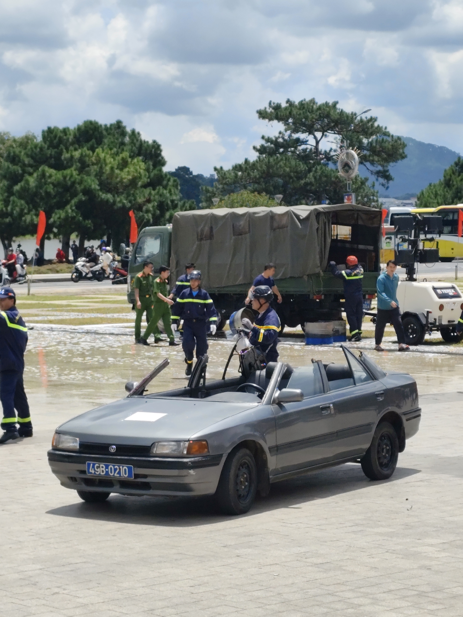 In Da Lat, Vietnam, on a cloudy day, a group of individuals in uniform are gathered near a gray convertible car. The car, identified by its license plate "49B-0210," is parked on a paved surface that appears to be wet and possibly flooded. Behind the car, a large military-style truck with a tarp-covered cargo area is visible. Several people in dark blue and green uniforms are standing or walking around the truck and a white generator trailer. One person in a dark blue uniform with yellow accents is standing in front of the car, appearing to be observing the scene. In the background, trees and a few orange flags are present. The overall atmosphere suggests an organized event or a response to an incident.