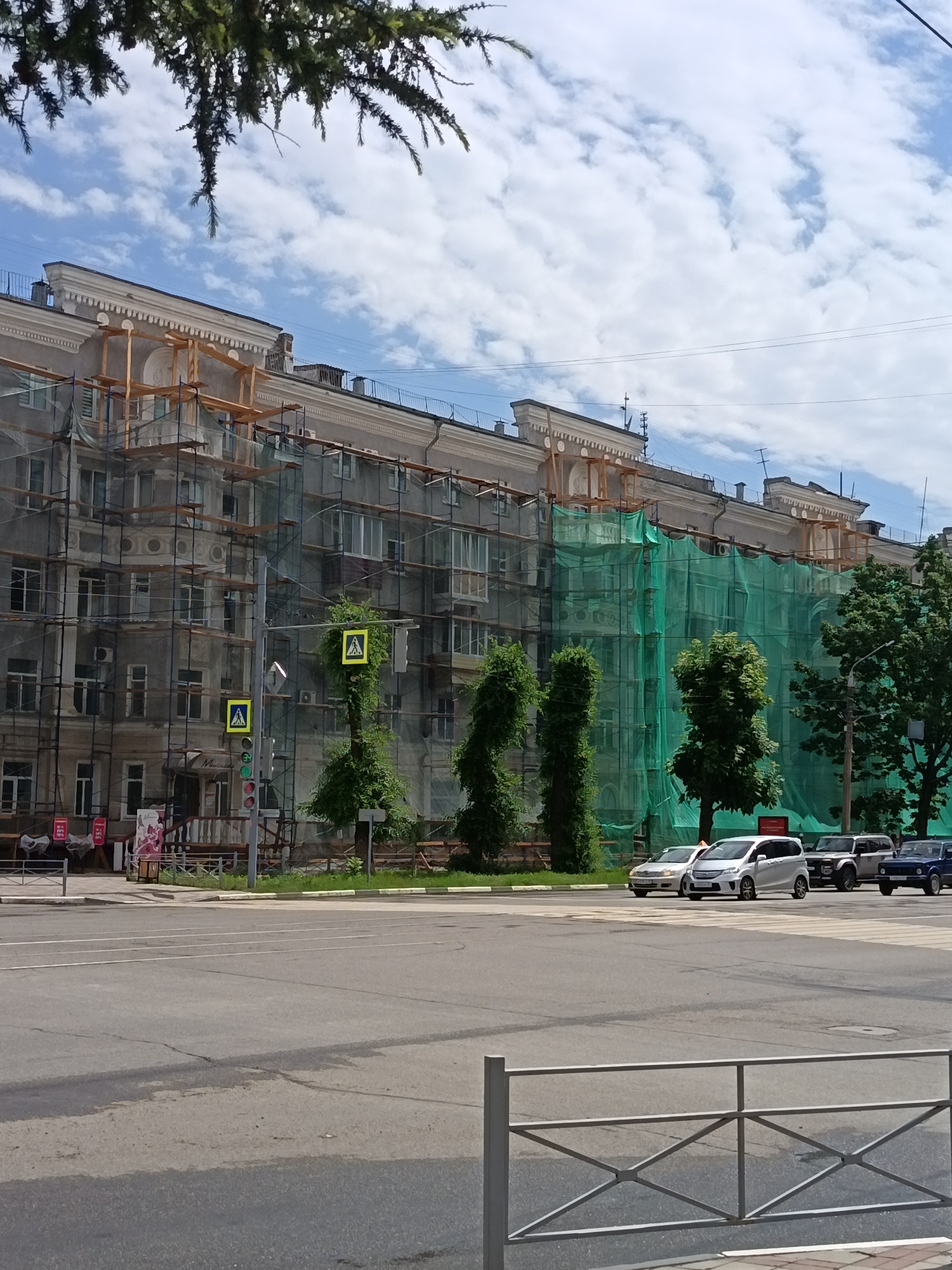 In Komsomolsk-on-Amur, Russia, an apartment building undergoing renovation is visible, covered in scaffolding and green netting. The building has multiple stories with visible windows and balconies. In the foreground, a street with a pedestrian crossing is shown, with cars driving on it. Traffic lights and pedestrian crossing signs are present on the side of the road. Trees and greenery surround the building and the street. The sky is partly cloudy.