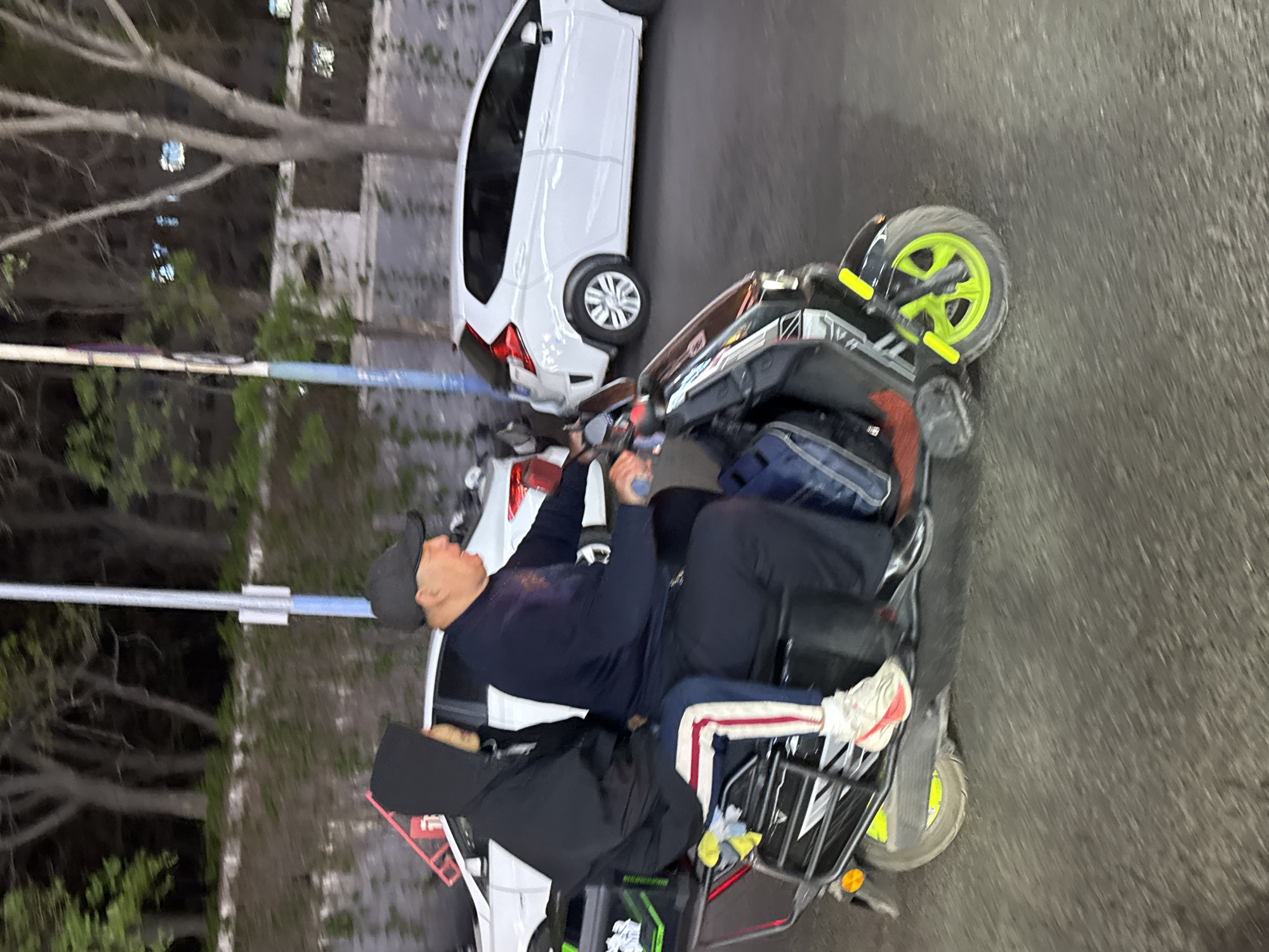 This nocturnal image, captured on a street in Zhongtun, China, depicts a three-wheeled electric scooter carrying two people. The scene is set at night, illuminated by artificial lighting from unseen sources and reflections from distant buildings.
The primary subject is the electric scooter, driven by a man wearing a dark cap, a dark long-sleeved shirt, and dark pants. His hands are on the handlebars, indicating he is operating the vehicle. Behind him, a second individual, partially obscured, is seated as a passenger, wearing dark clothing and a black hooded jacket. The scooter features a striking neon green front wheel with yellow accents on its fender, and a metal rack on the rear, upon which a blue bag is visible.
Behind the scooter, a white compact car, resembling a Honda Fit or similar model, is either parked or traveling on the paved road. The background consists of dark trees, some with bare branches and others with green foliage, suggesting an urban or suburban environment. Blurred buildings with illuminated windows are visible in the distance. A light-colored horizontal barrier or pole stretches across the middle ground. The overall mood is calm, indicative of nighttime activity in the area. No discernible text is visible on any objects.