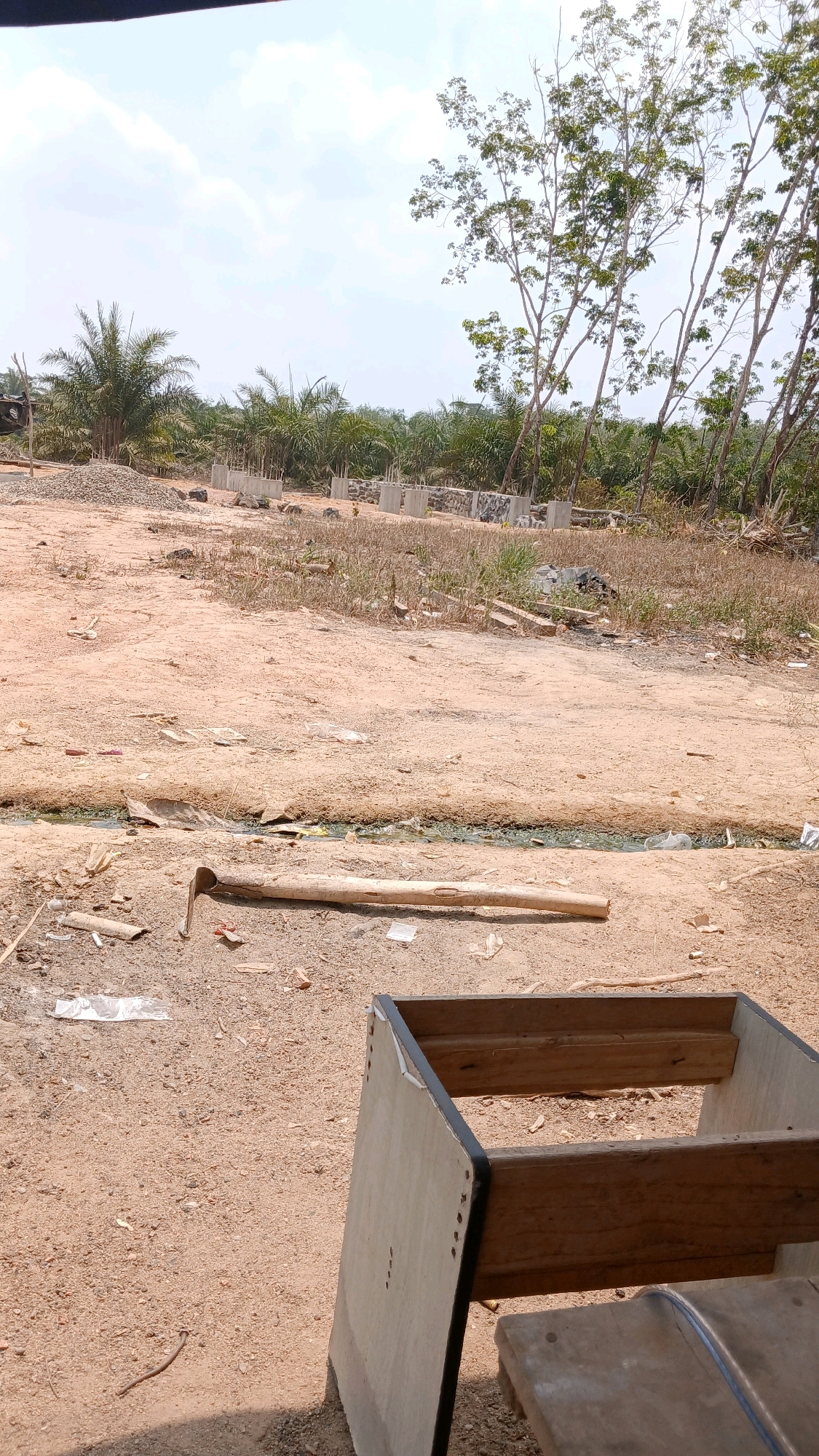 The image depicts an outdoor scene in Adi Luhur, Indonesia, likely a construction site or undeveloped land. The foreground features a dirt ground with scattered debris, including plastic bags, wood scraps, and a hoe lying horizontally across the mid-ground. Further back, a small water channel is visible, also containing debris. Beyond the channel, the ground is covered in dry grass and more scattered materials. In the background, concrete pillars and foundations are erected, indicating ongoing construction. Palm trees and other deciduous trees form a dense background against a bright, cloudy sky. In the lower right corner, a partially visible wooden structure, possibly a bench or part of a construction frame, is present. There are no people visible in the image.