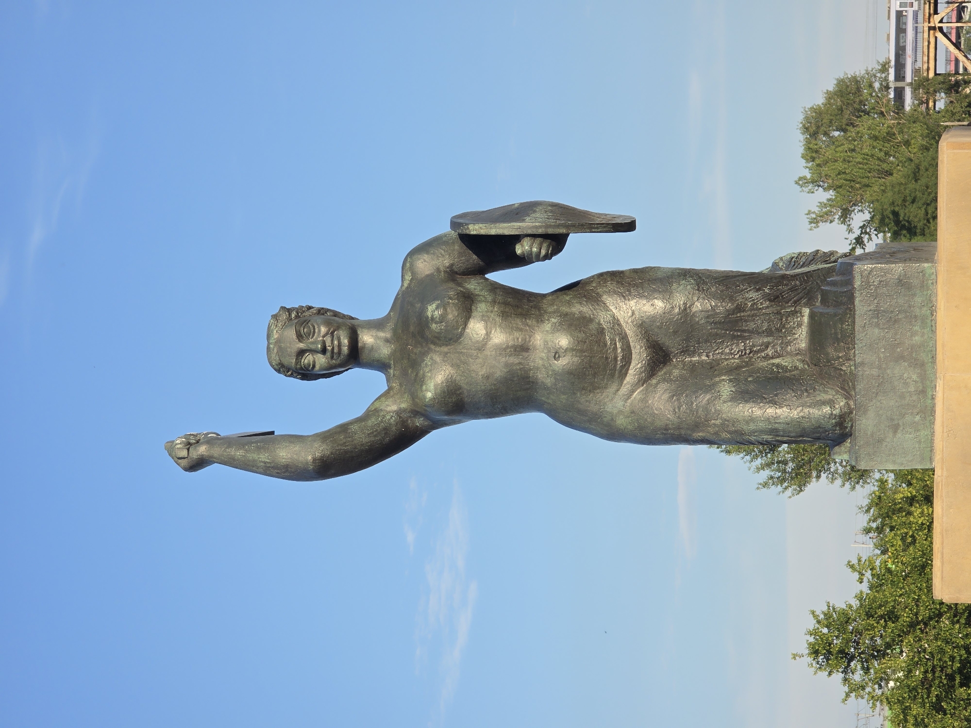 The image shows a bronze statue of a woman holding a sword in her raised right hand and a shield in her left. The statue is positioned in front of a clear blue sky with a few wispy clouds. Lush green trees are visible in the background, along with a concrete base supporting the statue and hints of urban infrastructure. This statue is the Warsaw Mermaid (Syrenka Warszawska), a prominent symbol of the city of Warsaw, Poland, located on the bank of the Vistula River in the Old Town.