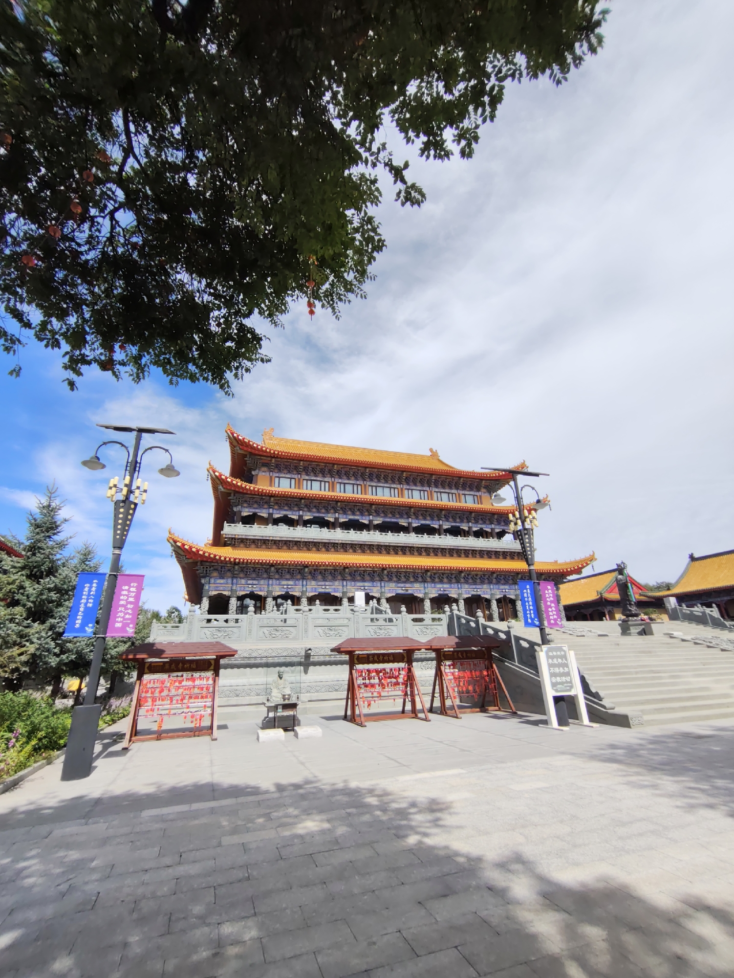 The image depicts a large, multi-tiered Buddhist temple with distinctive orange tiled roofs and ornate blue and gold detailing on its facade. The temple is constructed from grey stone and sits atop a wide set of stairs. In the foreground, a paved courtyard is visible, with shadows cast by trees and possibly a camera support. To the left of the temple, a tall lamppost stands with two lights. Hanging from the lamppost are several colorful banners, one blue and one purple, with Chinese characters. Adjacent to the lamppost and banners are two red wooden structures, each filled with numerous red tags or ribbons. A few evergreen trees are also present to the left of the temple. In the background, other temple structures with orange roofs are visible. The sky is blue with scattered white clouds. The scene appears to be a daytime shot under good weather conditions. This location is the Daqing City Buddhist Temple in Daqing, China.