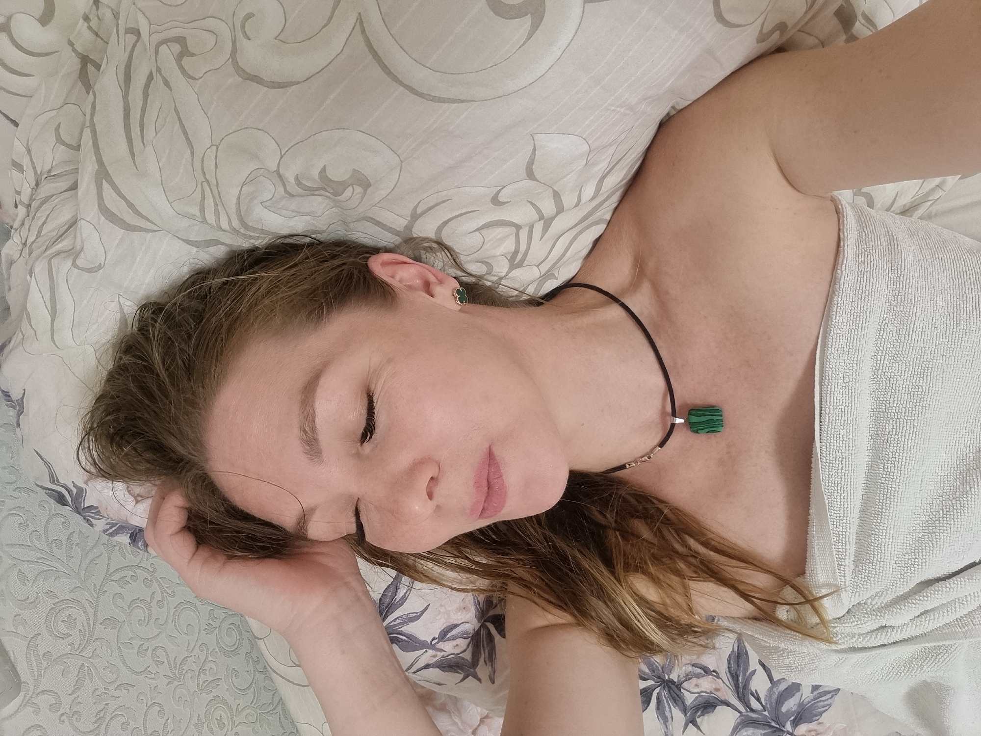 An overhead shot captures a woman lying peacefully on a bed indoors, likely in Chaikovsky, Russia. She appears to be sleeping or resting, with her eyes gently closed and a serene expression. Her head is tilted slightly to her left, resting on a pillow.
The woman has light brown or auburn hair, which is casually spread around her head. She has fair skin. In her visible right ear, she wears a small, dark green, clover-shaped stud earring with a gold-toned edge. Around her neck is a black cord necklace featuring a square green pendant, which displays natural striped patterns, characteristic of malachite. Her right hand is positioned gently near her head, with her fingers slightly curled.
She is partially covered by a white, textured fabric, resembling a towel or a light blanket, draped over her upper body. The bedding consists of a light-colored pillow adorned with a subtle, greyish abstract swirling pattern. Beneath her, the bed sheet or duvet features a floral design with light blue-grey leaves and delicate pink or white flowers. The soft, even lighting suggests an indoor setting. There is no visible text in the image.