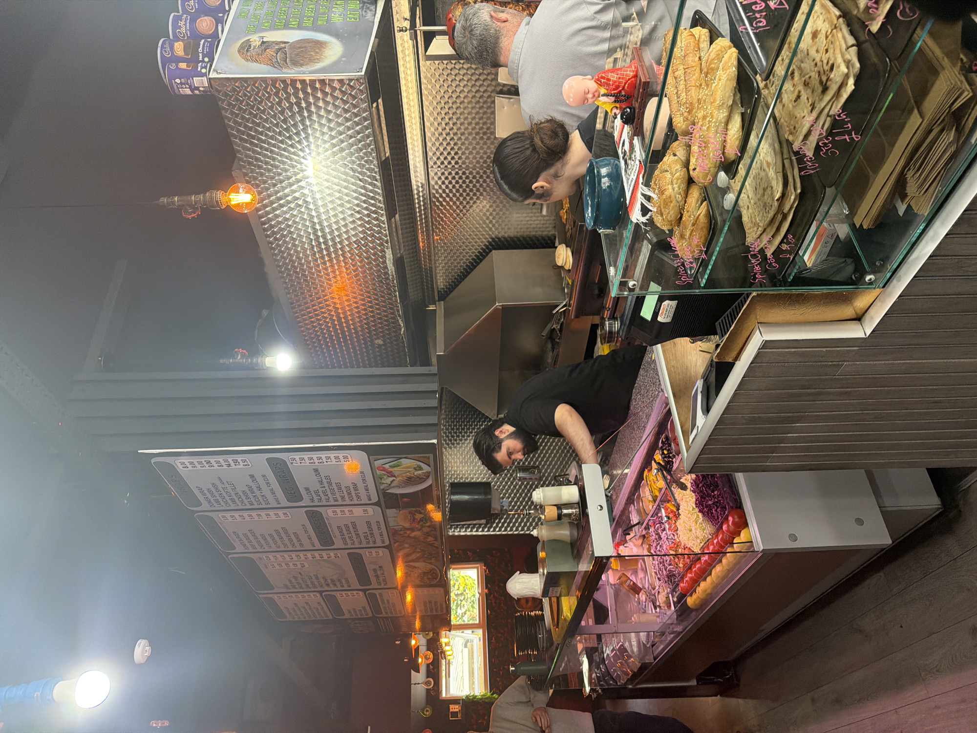 The image depicts the interior of a food establishment, possibly in London, United Kingdom. There are several people inside. A man wearing a black shirt and face covering is seen preparing food behind a glass display case filled with various ingredients such as salads and vegetables. To the right, behind a glass display case, are bread products like pastries and flatbreads. Customers are standing near the food preparation area. Overhead, a menu board lists various food items and prices. The environment suggests a casual dining setting.