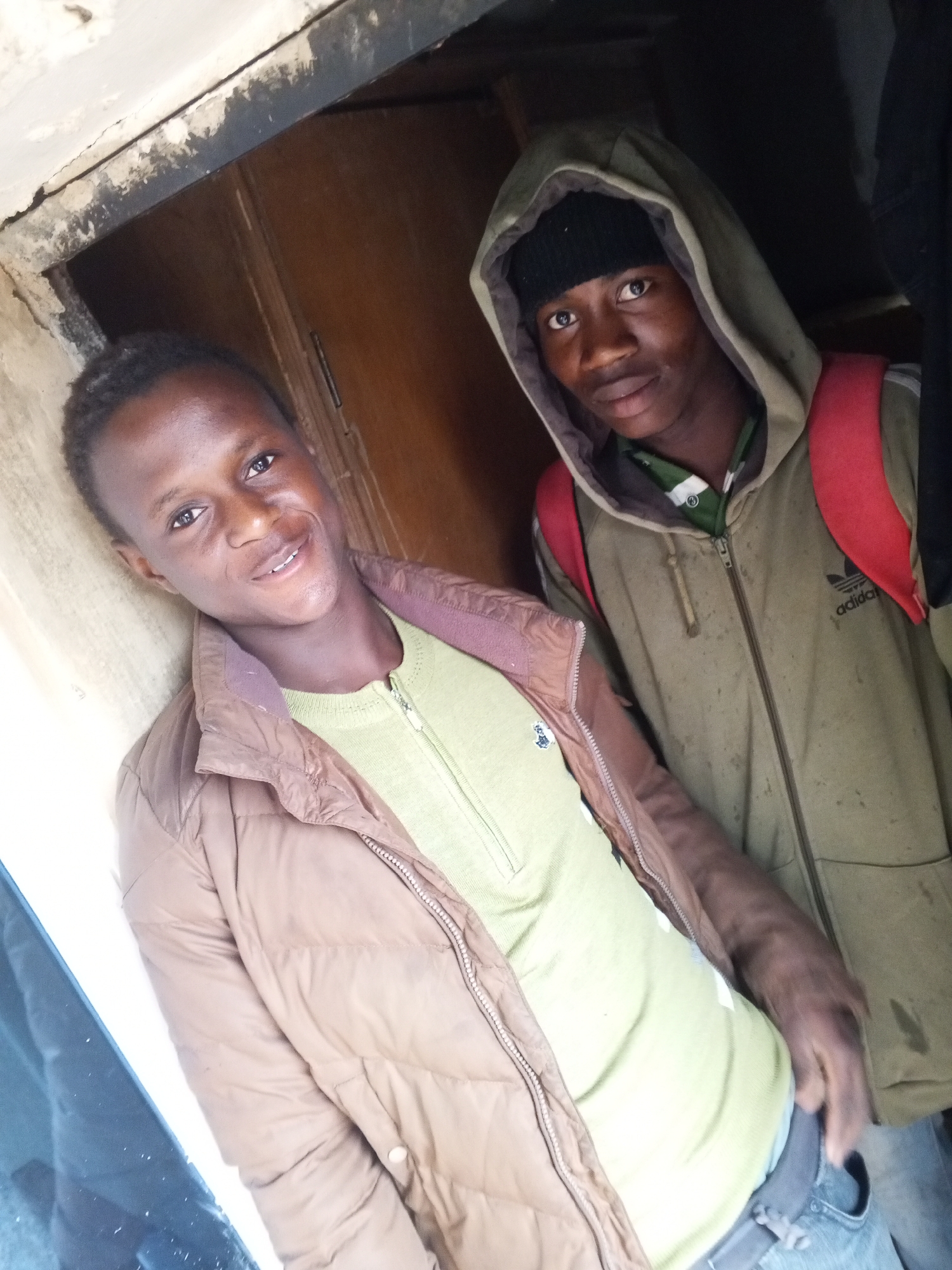 Two young men are posing for a photograph in what appears to be a doorway or narrow indoor space. The image is taken from a slightly low angle, tilted upwards and to the right.
On the left, a man with short, dark hair is smiling broadly, revealing his teeth. He is wearing a light green, long-sleeved shirt with a short zipper at the neck, layered under a brown puffer jacket. His right arm is slightly bent, and he is leaning against a light-colored, possibly stucco or painted wall that shows some wear and tear.
On the right, another man, looking directly at the camera with a neutral expression, is wearing an olive-green hooded jacket with the hood up, covering a black beanie. The jacket has a full zipper and an "adidas" logo visible on the right chest area. Red backpack straps are visible over his shoulders. He is wearing a striped shirt underneath his jacket. His left hand is tucked into his jeans pocket.
The background behind both men is dark, suggesting a deep doorway or an unlit interior. The wall on the left side of the image has visible cracks and chipped paint or plaster. The lighting is brighter on the left side, illuminating the first man more, while the right side of the image is considerably darker. Both individuals are looking towards the camera, indicating they are aware of being photographed.