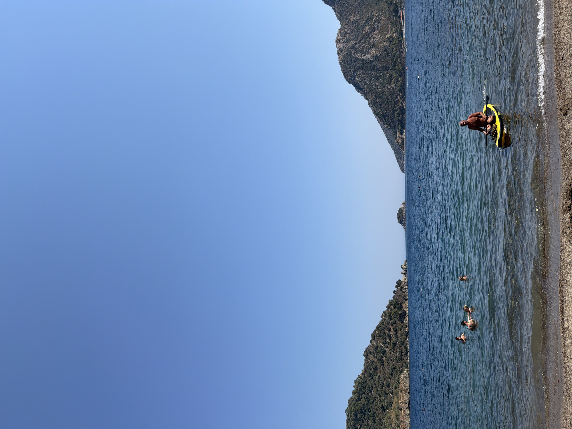 The image depicts a tranquil scene at a beach in Marmaris, Turkey. The foreground features a sandy shoreline with gentle waves lapping at the beach. The sea stretches out into the distance, a deep blue under a clear, cloudless sky.
In the water, several people are visible. Four individuals are swimming and interacting near the shore, appearing to be enjoying a leisurely swim. Further out, a man is seated on a yellow paddleboard, holding a paddle. He is facing the camera and appears to be resting or observing the surroundings.
The background is dominated by lush green hills that rise from the sea, characteristic of the coastal landscape of Marmaris. The hills are covered in dense vegetation and appear to be rocky. In the distance, a small island or rocky outcrop is visible, adding to the scenic beauty of the location. The overall impression is one of a peaceful summer day at a beautiful Mediterranean beach.
