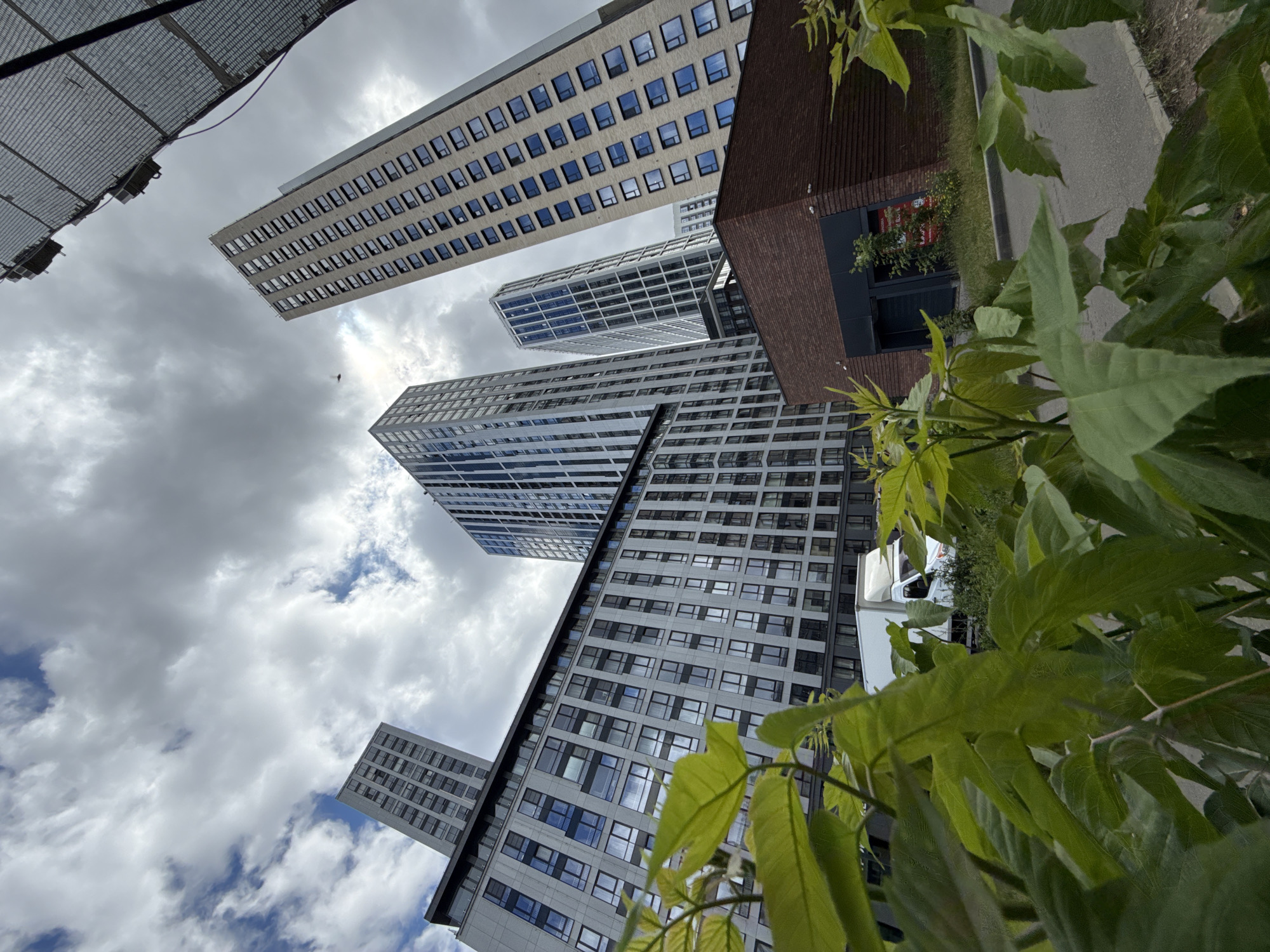 The image depicts a modern cityscape dominated by high-rise buildings under a cloudy sky. In the foreground, lush green foliage partially obscures the view. Several multi-story residential or office buildings with geometric facades and numerous windows are visible. One building in the mid-ground appears to be made of light-colored brick, while others feature glass and darker materials. A white van is parked on the street in front of one of the buildings. A low, brick-clad structure with a dark entrance is situated to the right of the image. The overall scene suggests a residential or mixed-use development within an urban environment. The presence of specific architectural styles and the white van, commonly used for deliveries, suggest an urban setting in Moscow, Russia, likely a newly developed area.